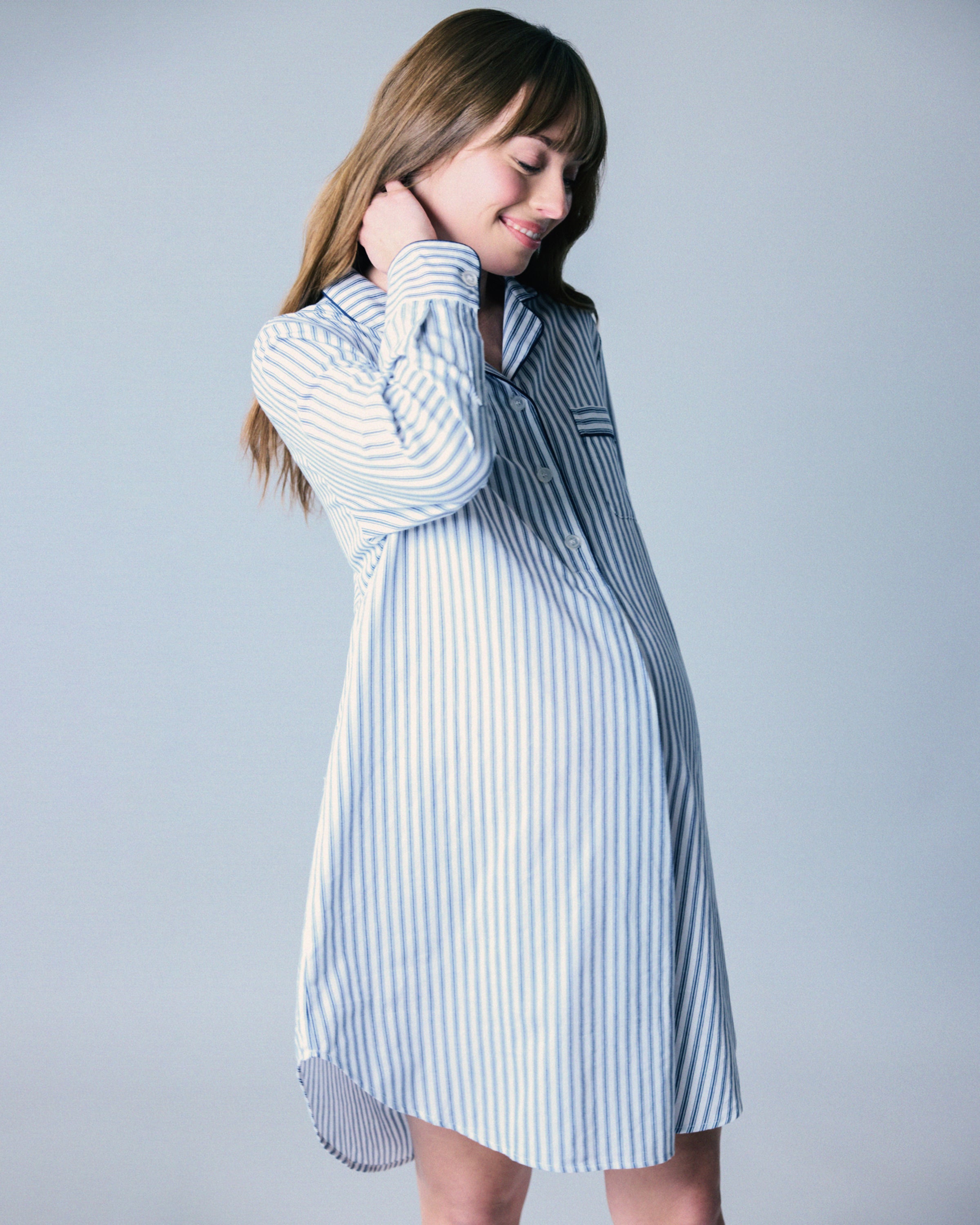 A pregnant woman with long brown hair gently touches her neck, wearing a luxurious Womens Twill Maternity Nightshirt in Navy French Ticking by Petite Plume. The soft cotton fabric envelops her comfortably as she smiles softly against a light gray background.