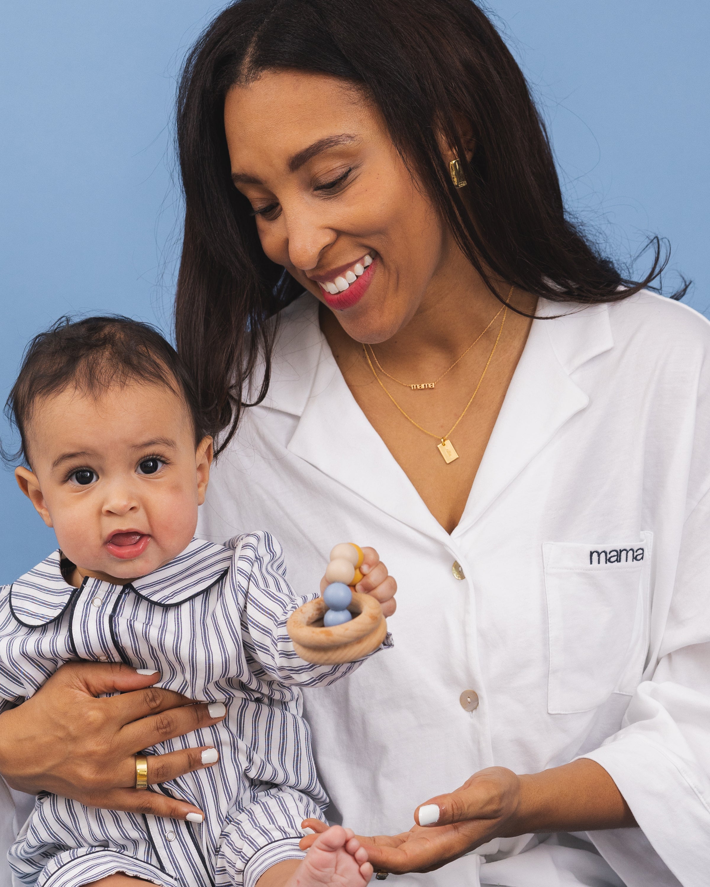 Cradling her baby, a woman smiles in Petite Plumes Olivia Wide Leg Pima Pajama Set in White, featuring Mama embroidery. The baby, dressed in stripes and holding a toy, adds to the tenderness of this scene against a light blue backdrop, epitomizing Mothers Day.