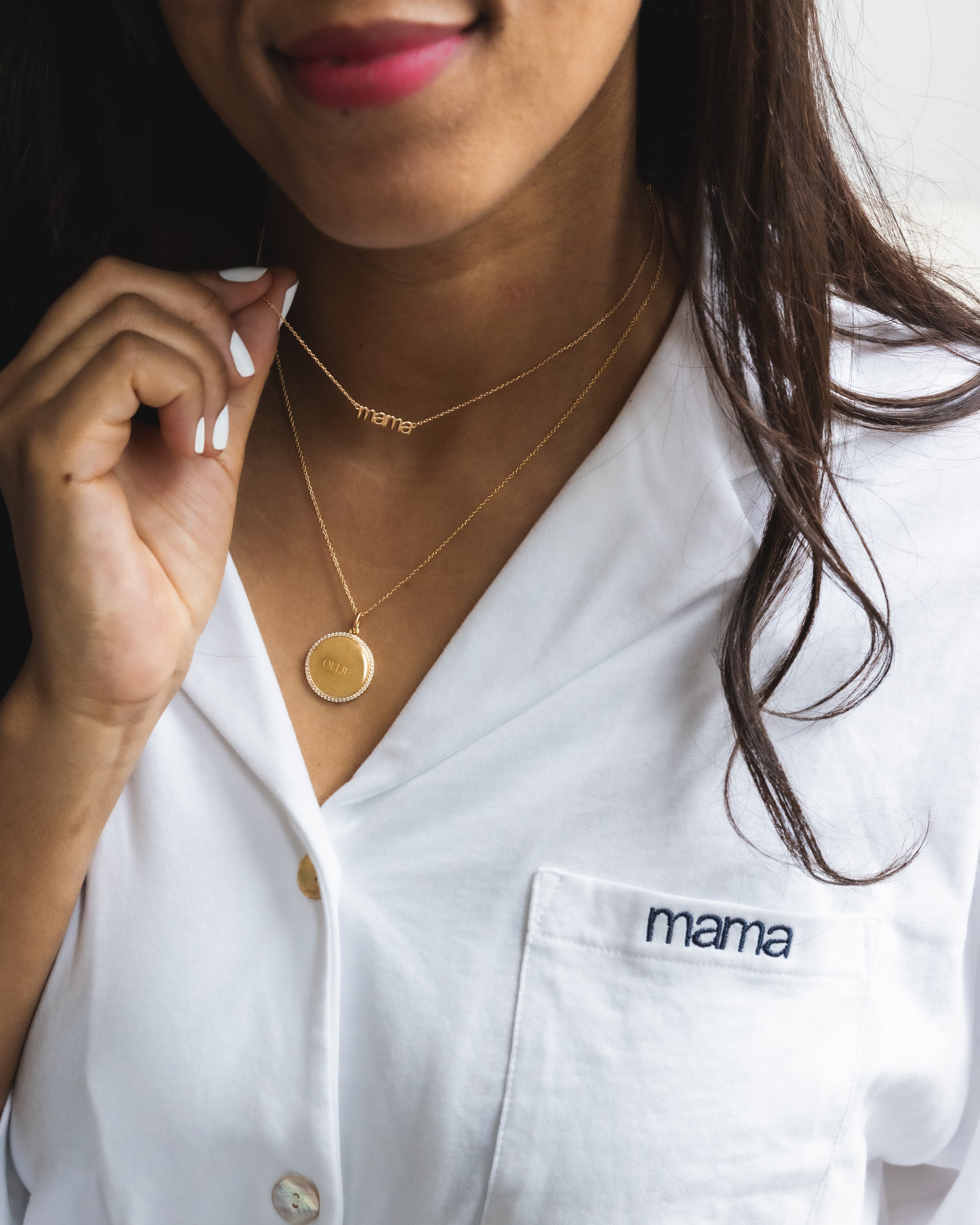 A woman models The Olivia Wide Leg Pima Pajama Set in White with Mama embroidery by Petite Plume, styled with two gold necklaces—one spelling mama and another with a pendant. Her white nails and long brown hair add to her elegance, while the set is crafted from luxurious Peruvian Pima cotton.