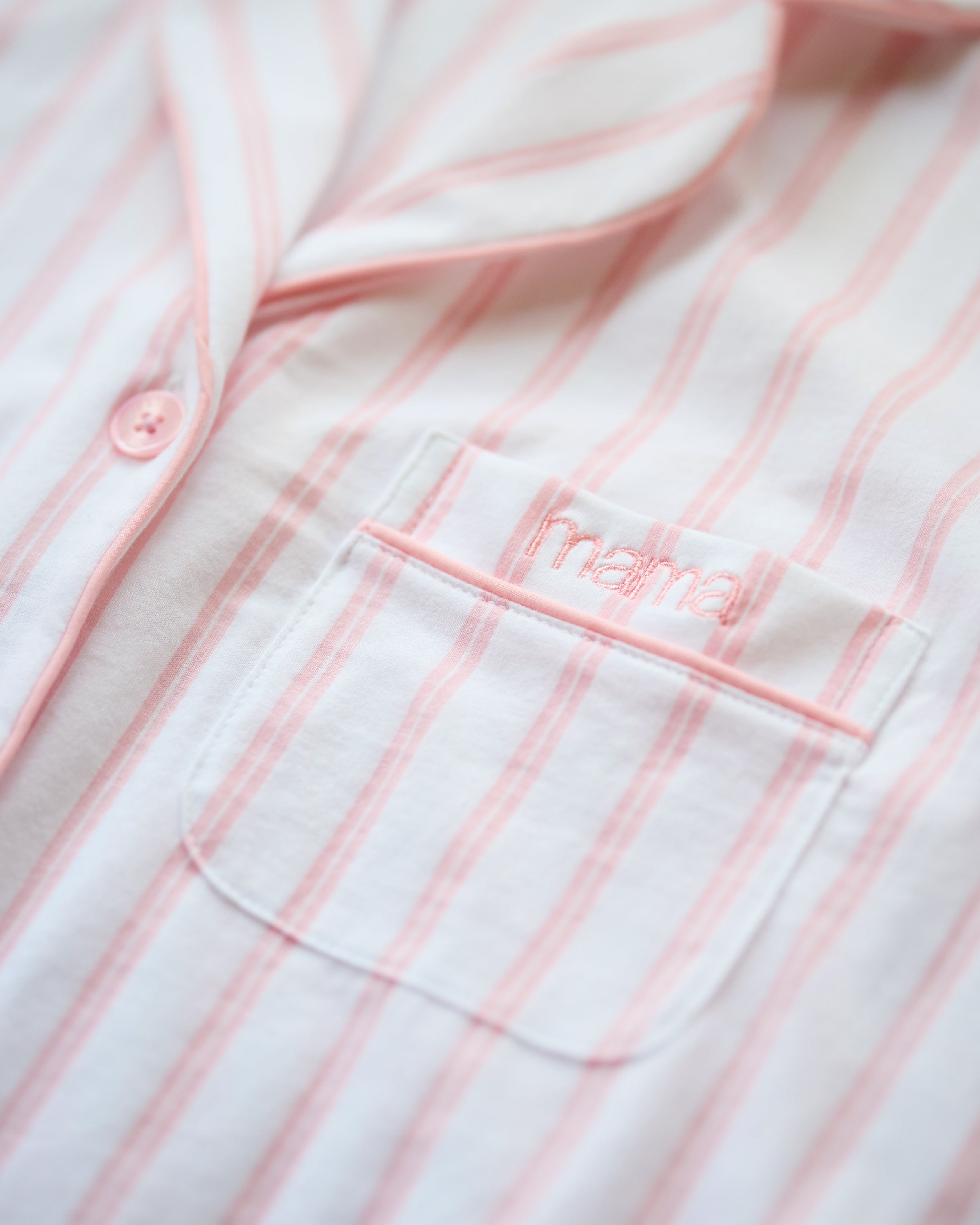 A close-up of luxurious fabric features Petite Plumes Womens Pima Pajama Set in Pink Stripe with Mama embroidered above the chest pocket. It includes a pink button and collar, perfect as an elegant Mothers Day edition.