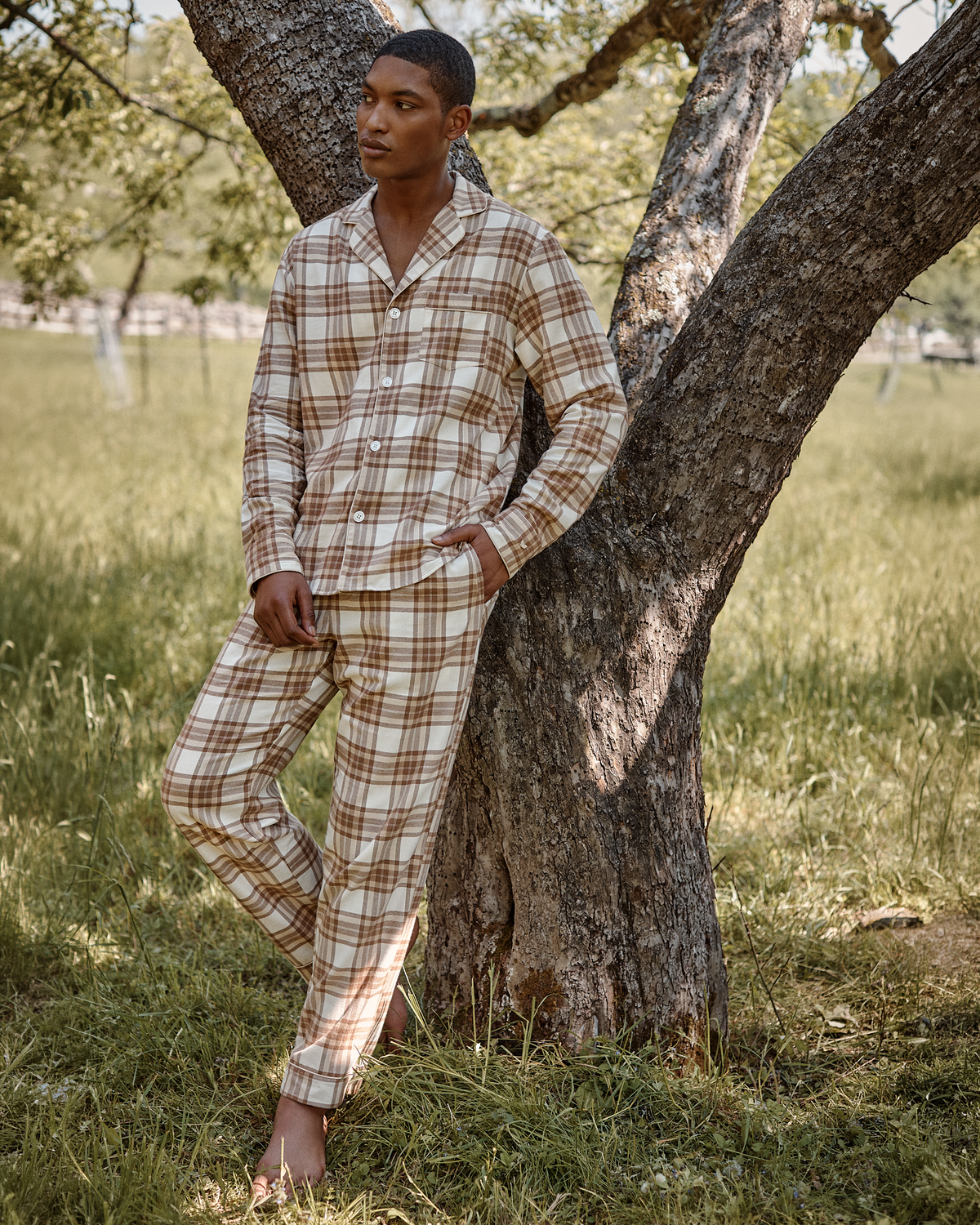 A person wearing the Men’s Twill Pajama Set in Cotswolds Tartan leans against a tree in a grassy outdoor setting on a sunny day.