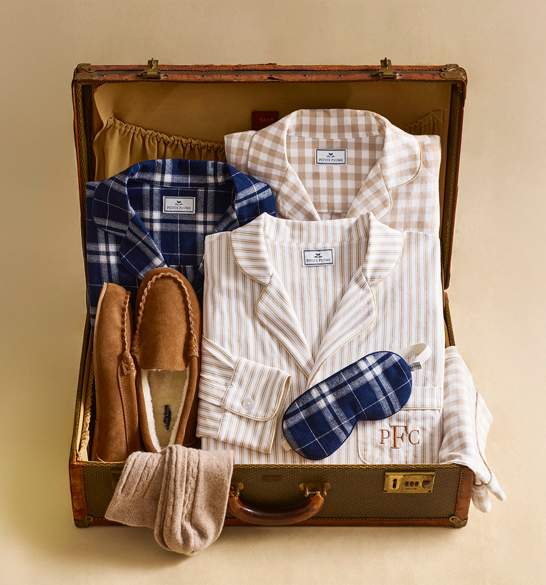 An open suitcase contains two sets of pajamas, one blue plaid and one beige striped, along with a matching striped sleep mask, tan slippers, and a pair of beige socks, all neatly arranged.