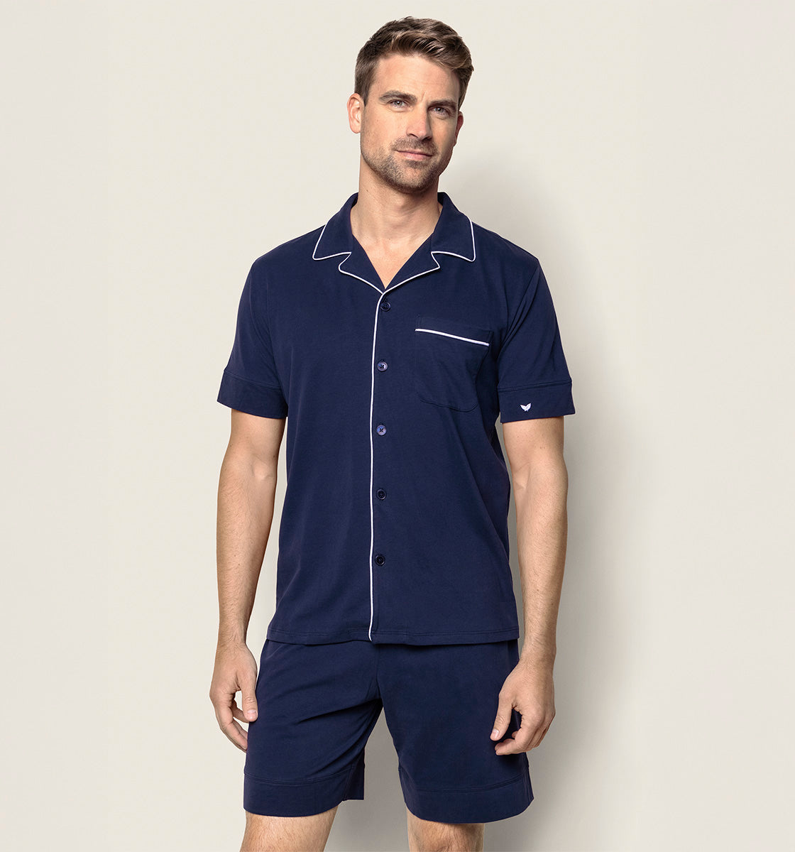 A man wearing a navy blue short-sleeve pajama set with white piping, including a button-up shirt with a chest pocket and matching shorts, stands against a plain light background.