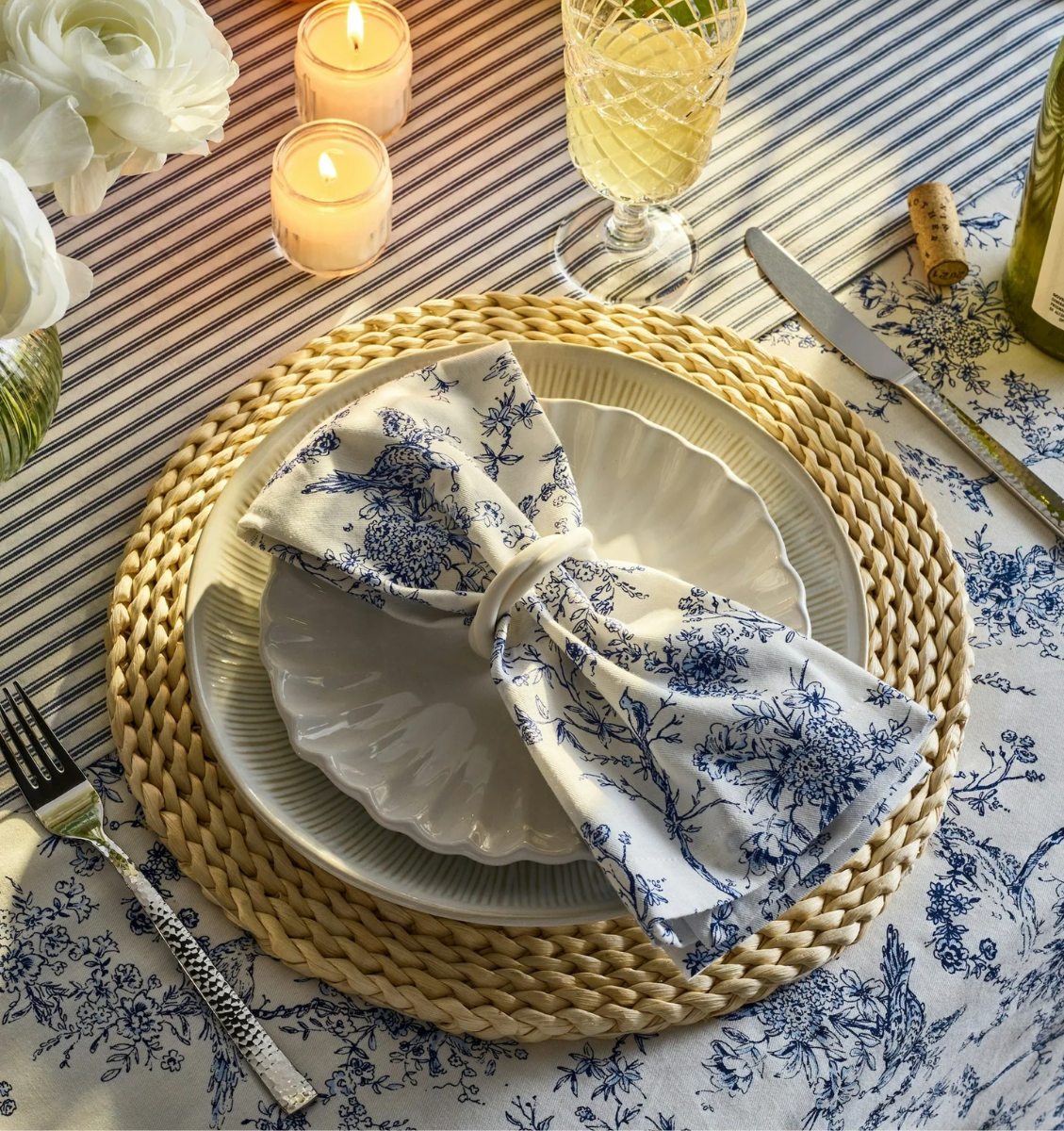 A table set with a woven placemat, scalloped white plates, a blue floral napkin shaped like a bow, a wine glass, candles, and floral tablecloth, creating an elegant, cozy dining atmosphere.
