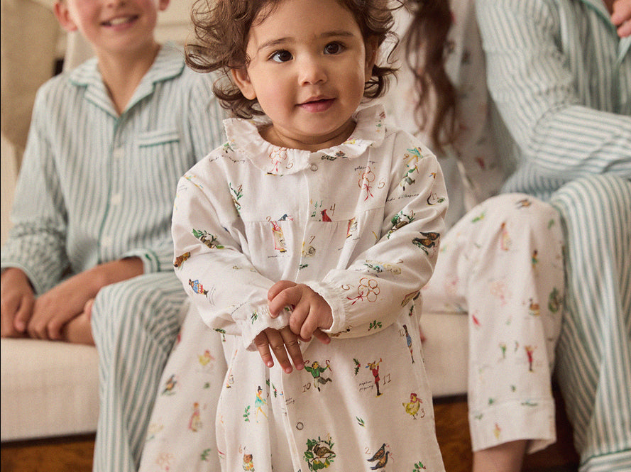 A toddler in patterned pajamas stands in front of adults and a child sitting on a couch, all wearing coordinating pajamas. The child looks at the camera with a gentle expression.