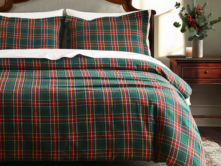 A neatly made bed with green, red, and white plaid bedding and matching pillows, next to a wooden nightstand with a vase of flowers and a lit candle.