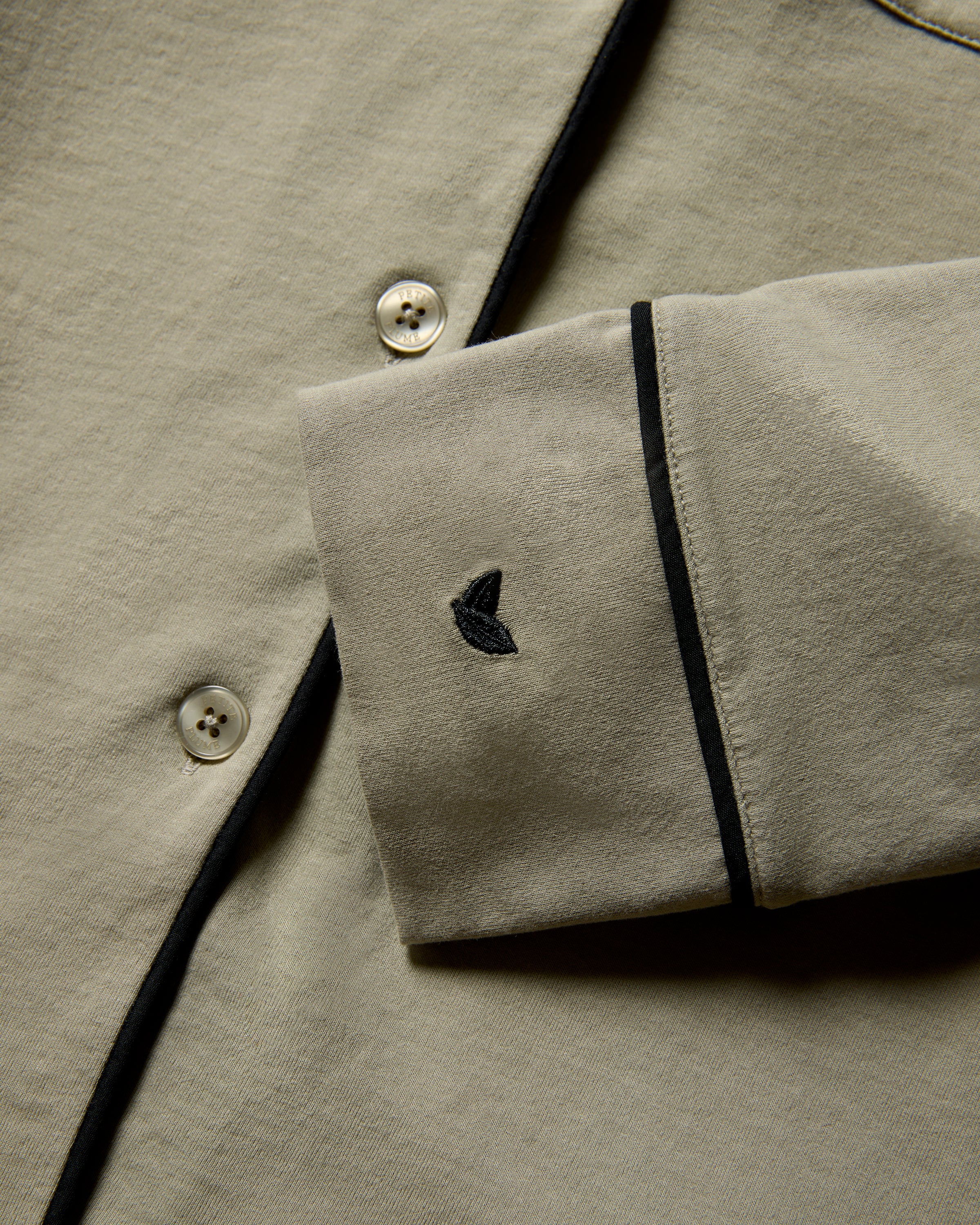 Close-up of a moss-colored shirt with black piping, made from plush Peruvian Pima cotton. The cuff sports a small embroidered logo in black. The shirt features white buttons and a smooth texture, part of the Petite Plume x Parachute Womens Pima Pajama Set.