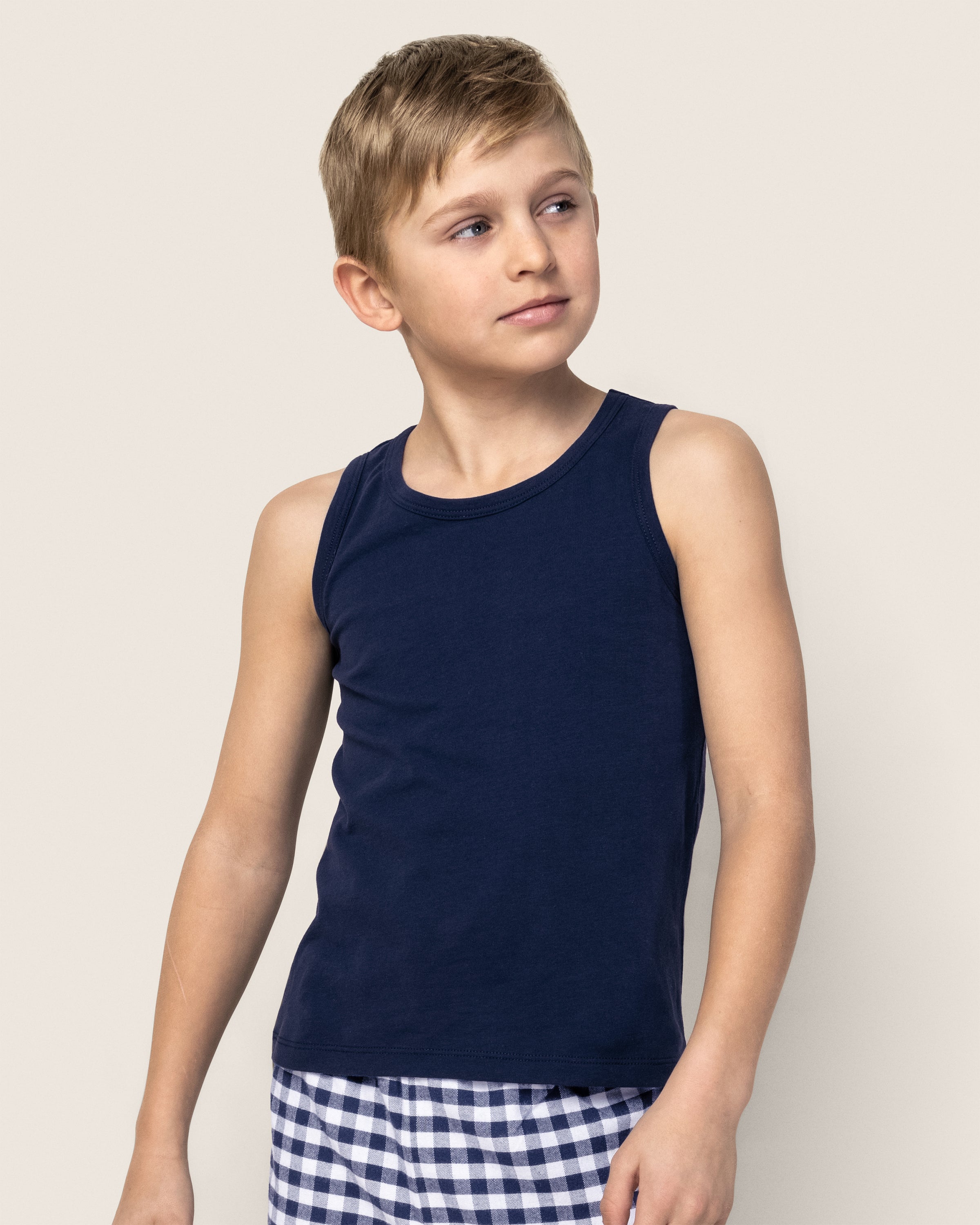 A young boy stands against a plain light background, looking to the side, wearing Petite Plumes Kids Pima Tank Top in navy blue and checkered shorts.