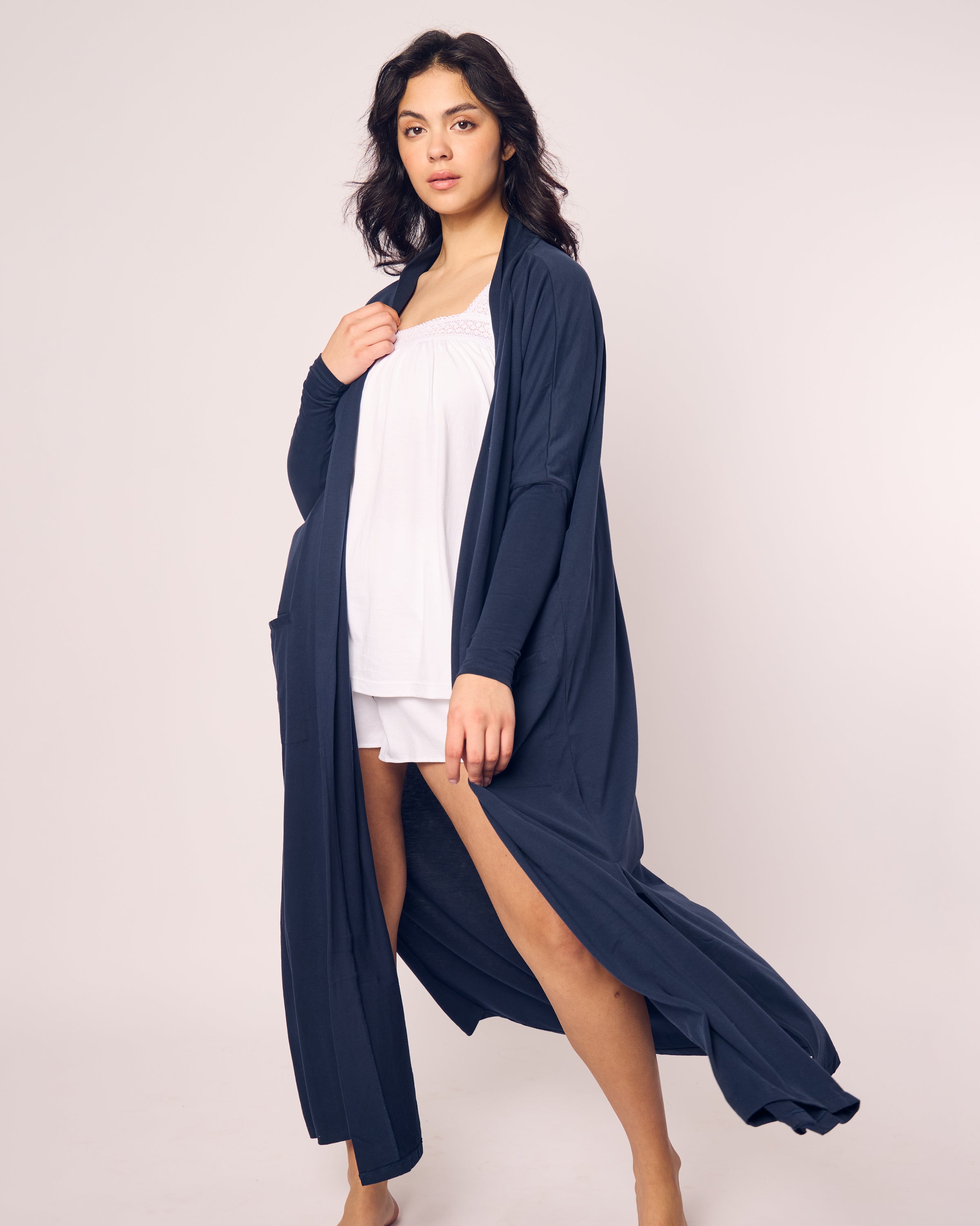 A person stands before the camera in Petite Plumes Womens Pima Duster in Navy, revealing luxury pajamas made from Peruvian Pima cotton underneath. The robe is open, and the background is plain and light-colored.
