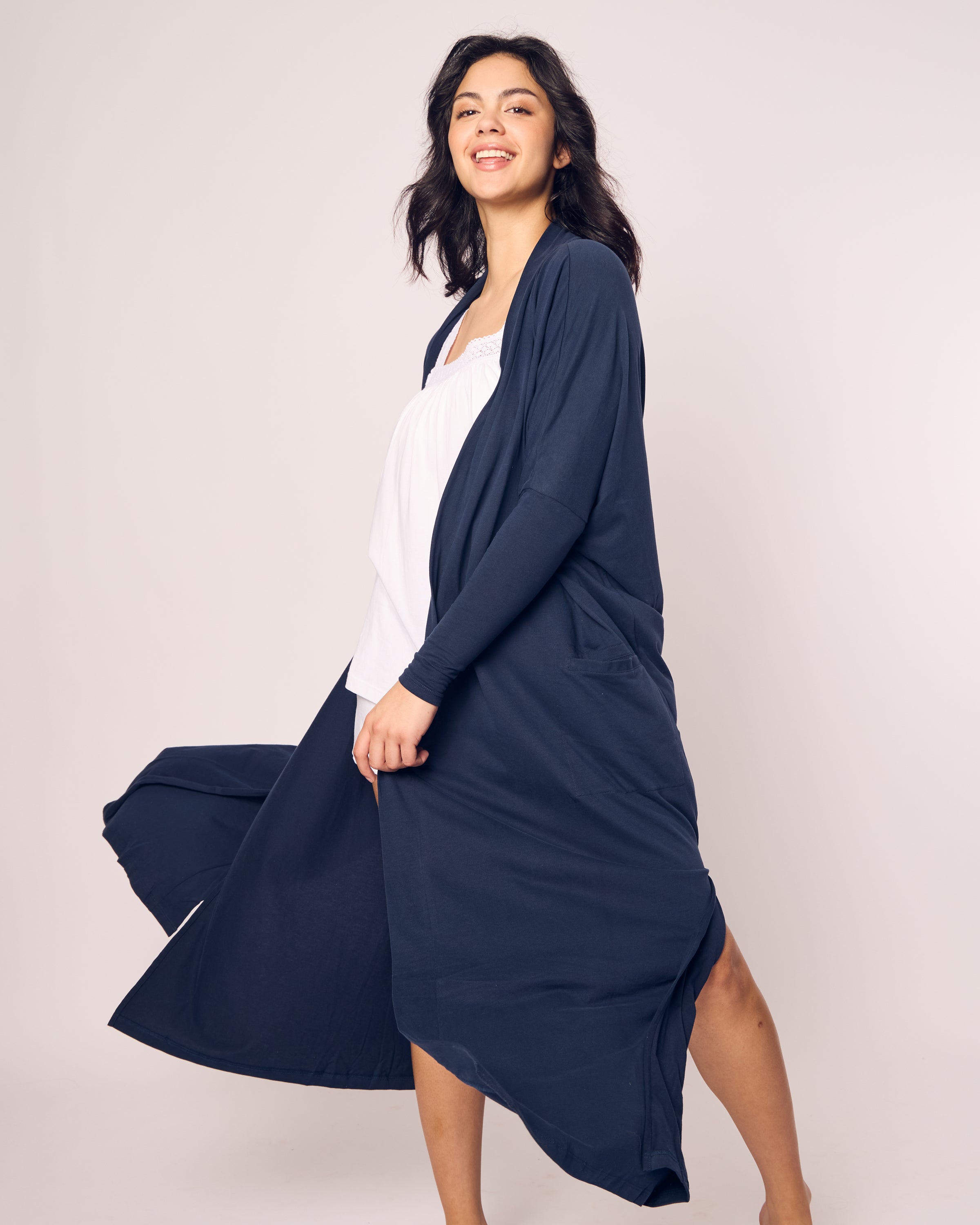 A person with long dark hair smiles while walking, wearing a flowing Navy Womens Pima Duster by Petite Plume over a white top. The duster flares out behind them, creating a sense of movement against a plain background.