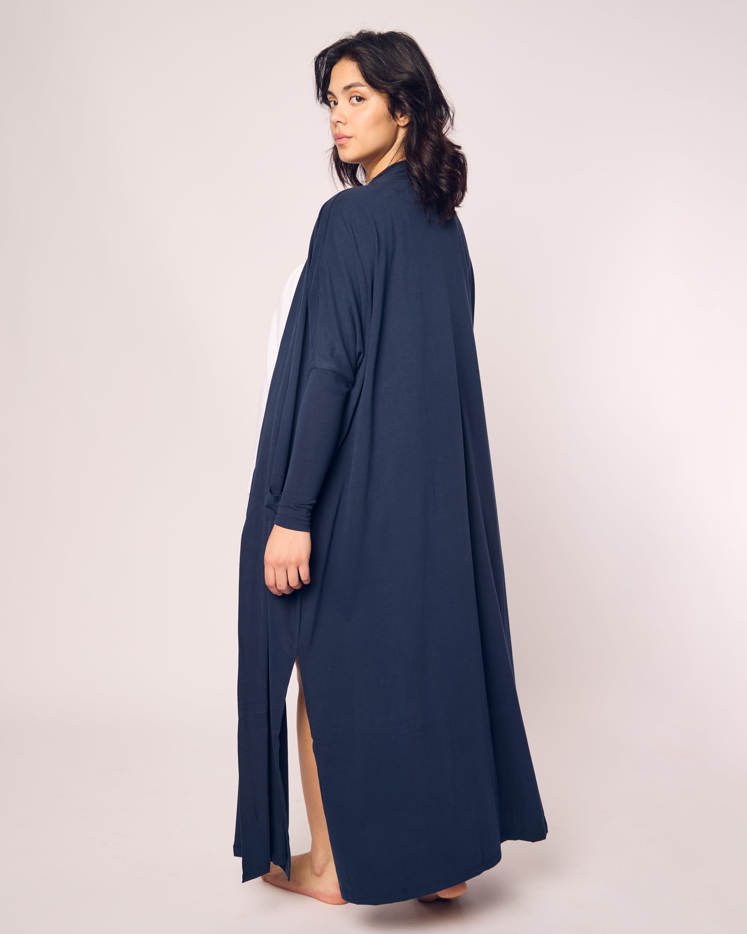 A person with dark hair stands barefoot wearing the luxurious Petite Plume Womens Pima Duster in Navy, made from breathable Peruvian Pima cotton. They look over their shoulder towards the camera against a plain, light-colored background.