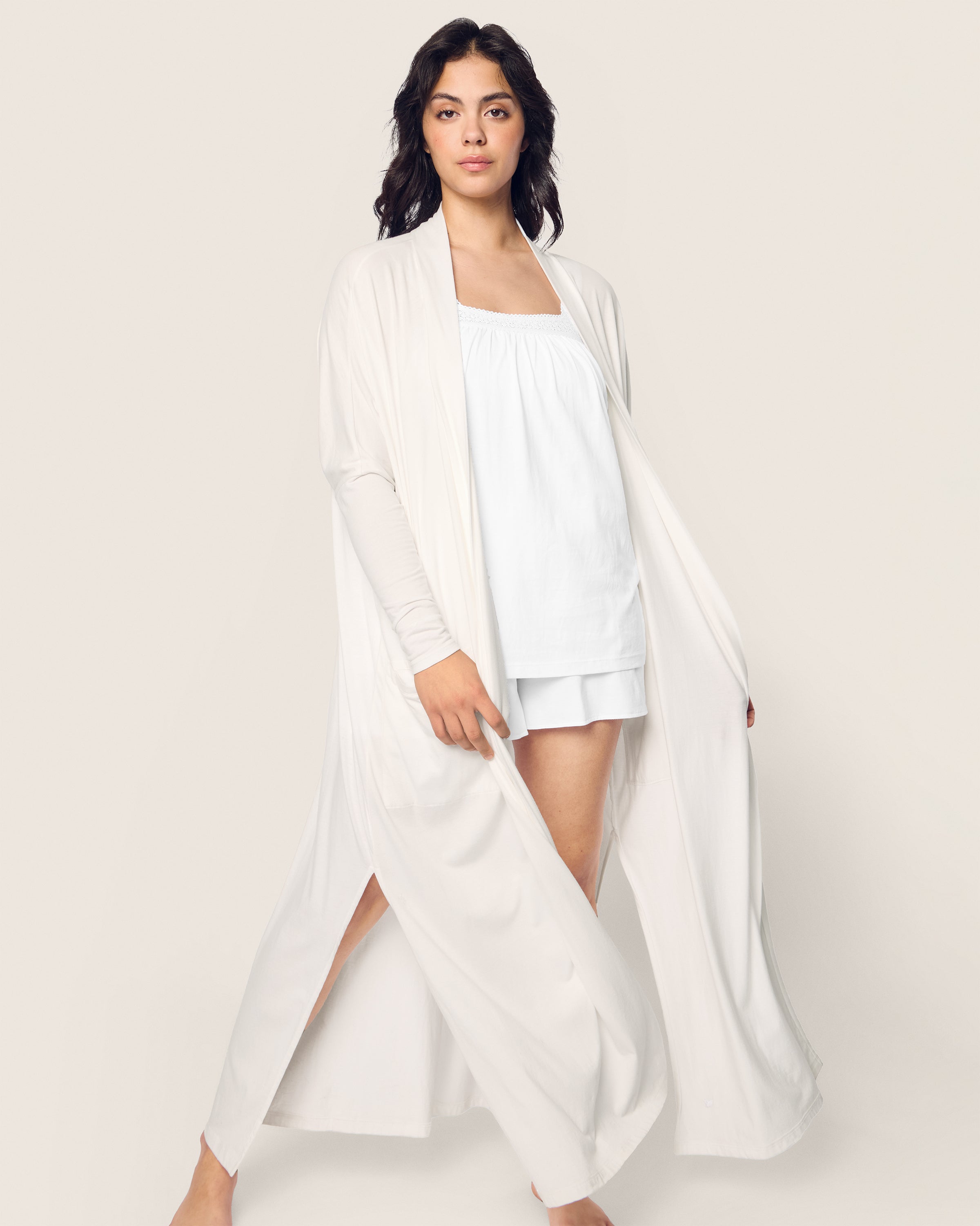 In front of a light neutral background, someone wears Petite Plumes Womens Pima Duster in White over luxurious pajamas made from Peruvian Pima cotton. Their shoulder-length dark hair frames their neutral expression, epitomizing the elegance and comfort of this breathable sleepwear.