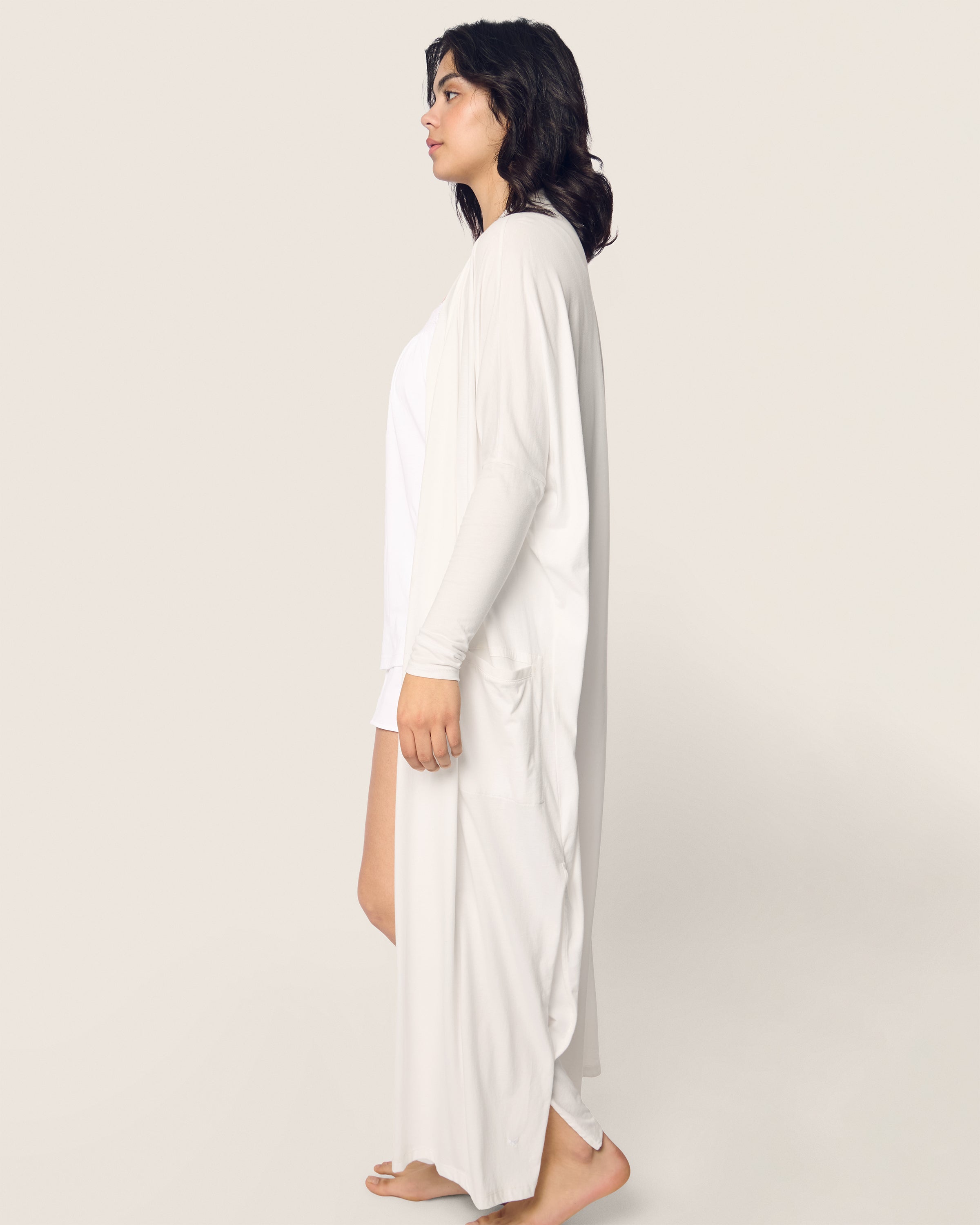 A woman with long dark hair stands barefoot in profile, wearing Petite Plumes Womens Pima Duster in White over luxurious Peruvian Pima cotton pajamas. The plain, light background enhances the tranquil elegance of her breathable sleepwear.