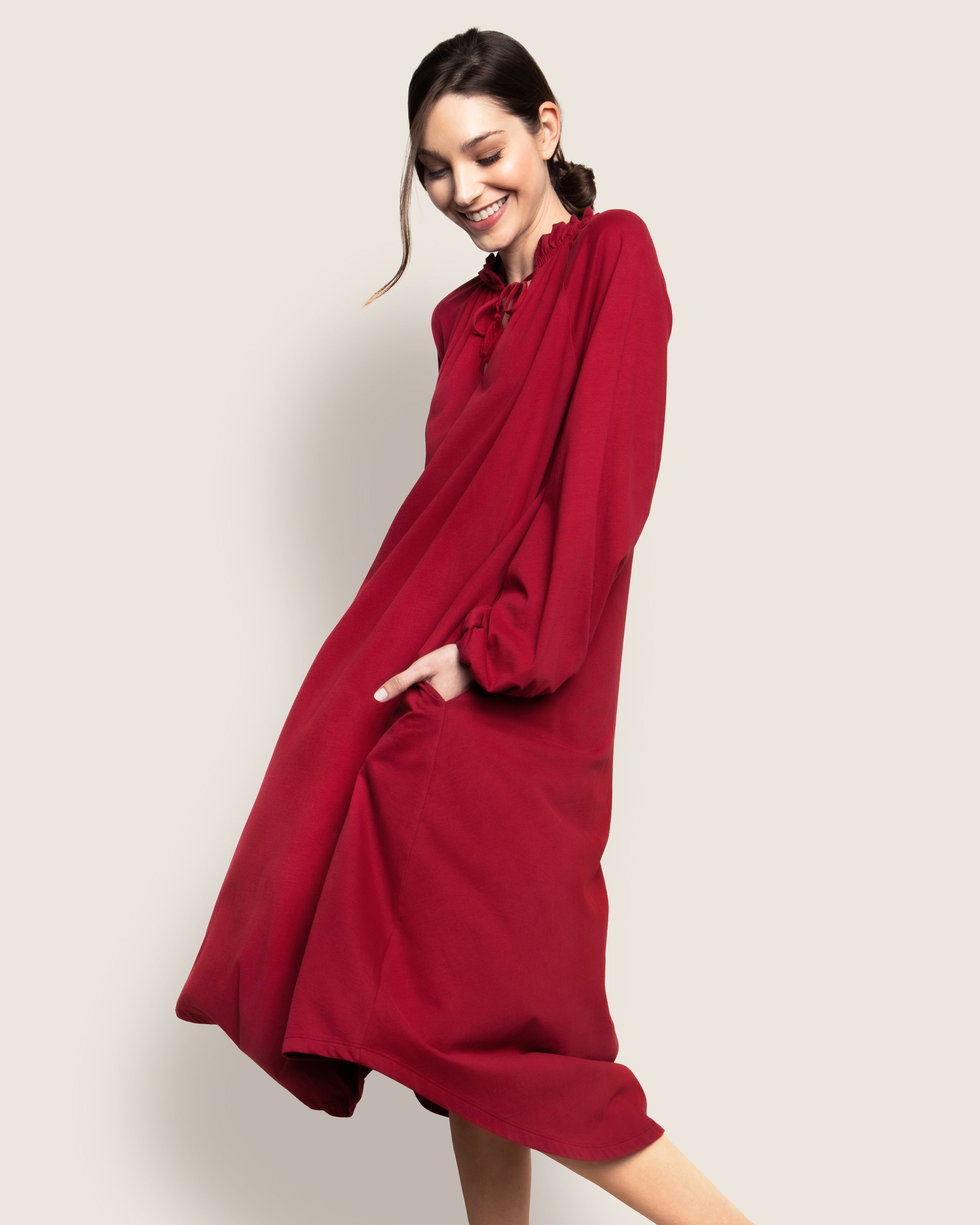 A person smiles while wearing the Petite Plume Womens Pima Garbo Nightgown in Bordeaux, a loose-fitting, long-sleeved gown reminiscent of a stylish nightgown. Crafted from luxurious Peruvian Pima cotton, it features a ruffled neckline and falls below the knees with their right hand in a pocket.