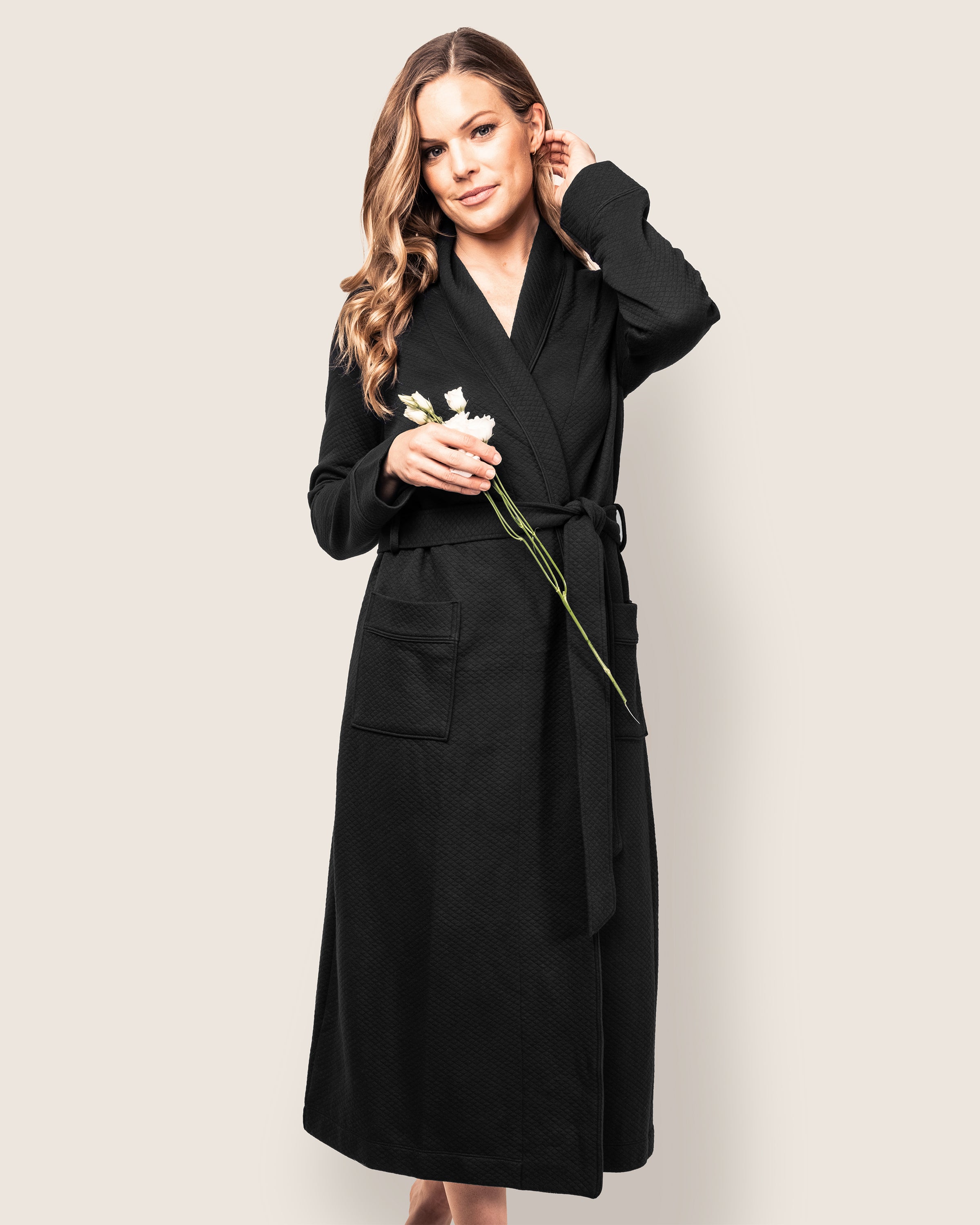 Against a light backdrop, a person wears the Womens Pima Ophelia Robe in Black Jacquard by Petite Plume. With their head tilted holding two white flowers, their other hand near their ear emphasizes the robes luxurious quilted design.