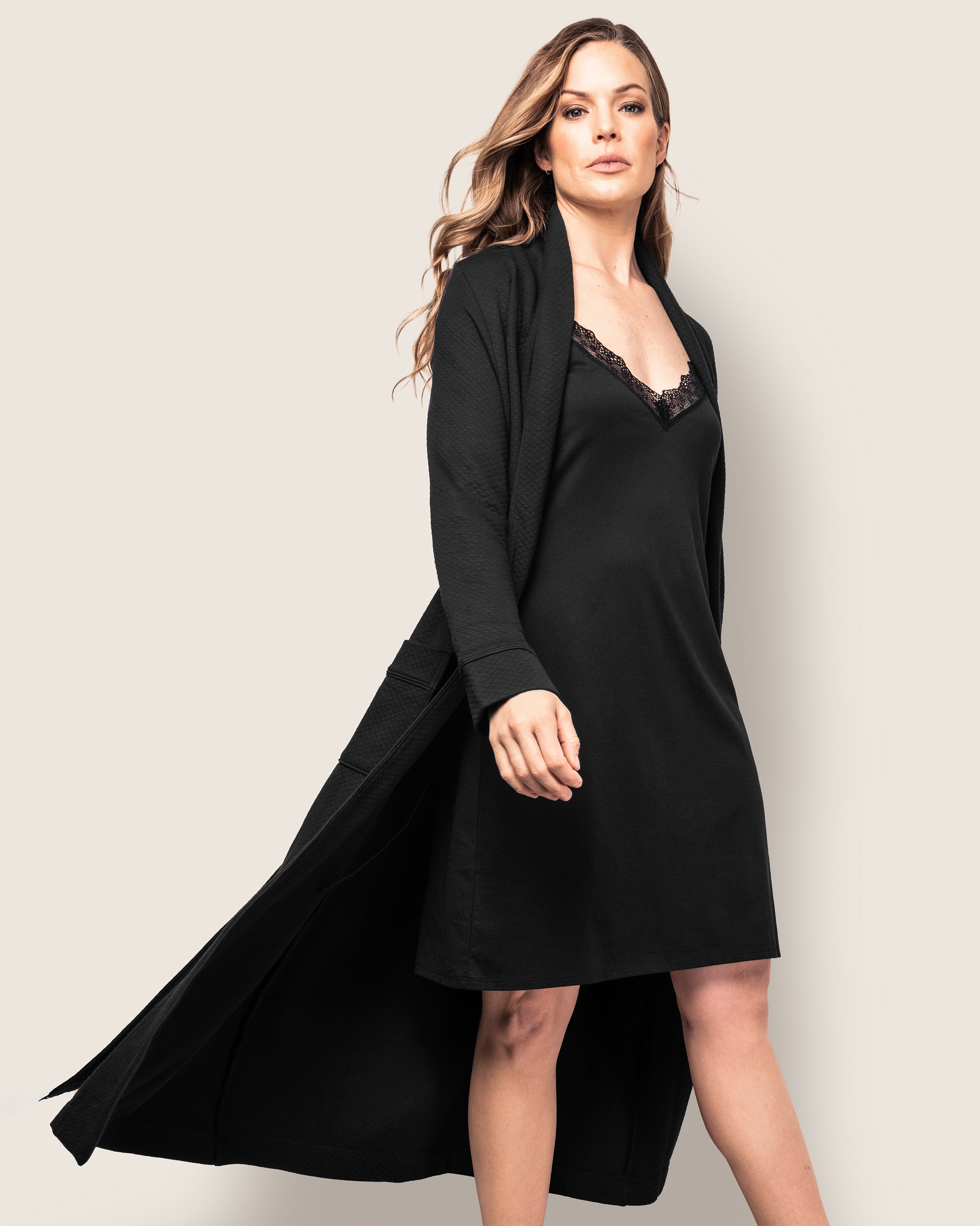 A person with long hair gracefully moves against a plain background, wearing a black dress featuring a lace neckline and the luxurious Petite Plume Womens Pima Ophelia Robe in Black Jacquard, its quilted effect flowing elegantly behind them.