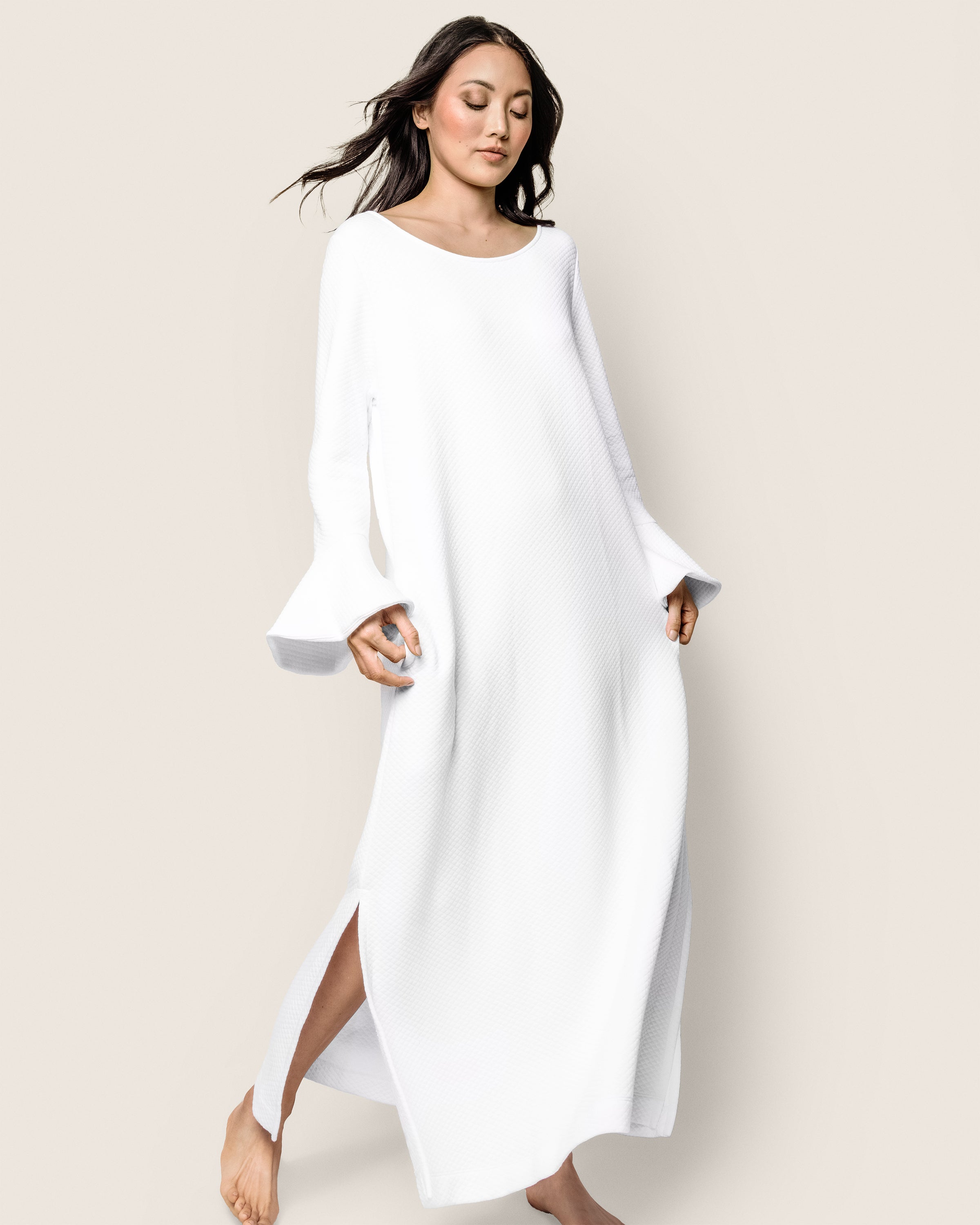 A woman wearing the Womens Pima Ophelia Nightgown in White Jacquard by Petite Plume walks barefoot against a light backdrop. Her dark hair streams behind her, and she gently holds the gowns bell sleeves with side slits, exuding serenity.
