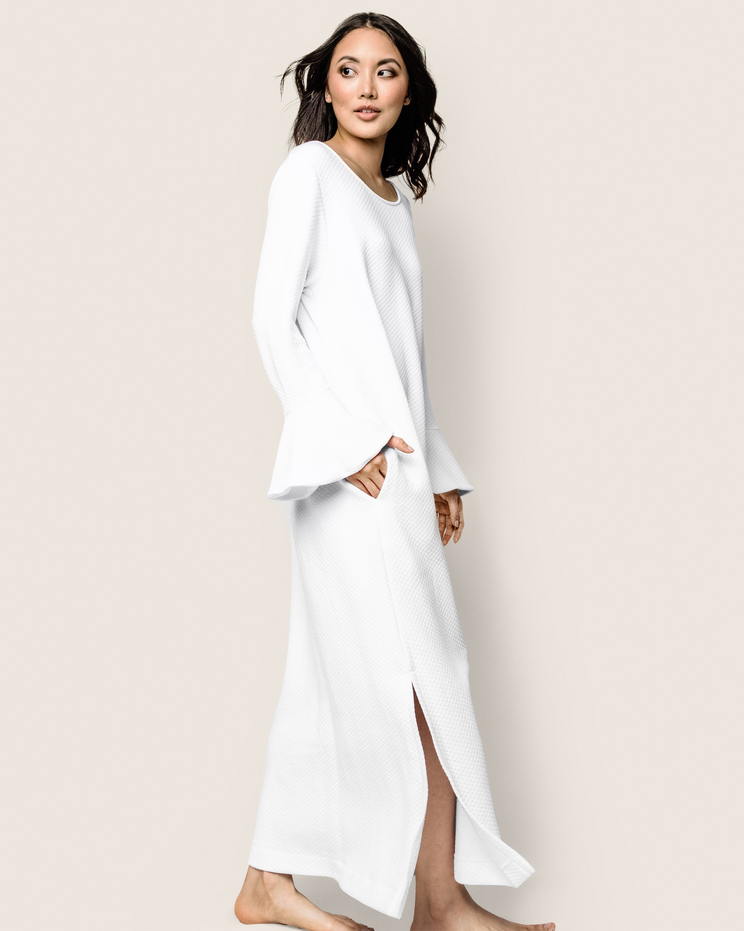 A woman models the Petite Plume Womens Pima Ophelia Nightgown in White Jacquard, barefoot against a plain backdrop. The nightgown, with long sleeves and a side slit near the hem, complements her loose dark hair and subtle smile as she gazes at the camera.