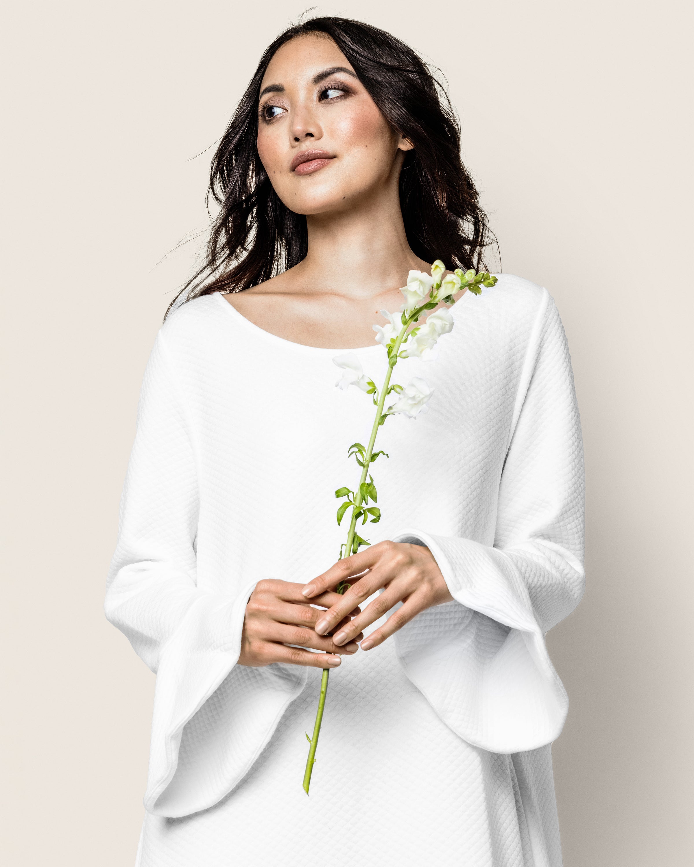 A woman in a Petite Plume Womens Pima Ophelia Nightgown in White Jacquard holds a single white flower, gazing slightly to the side against a light beige backdrop, her dark hair softly draping over her shoulders like Peruvian Pima cotton.