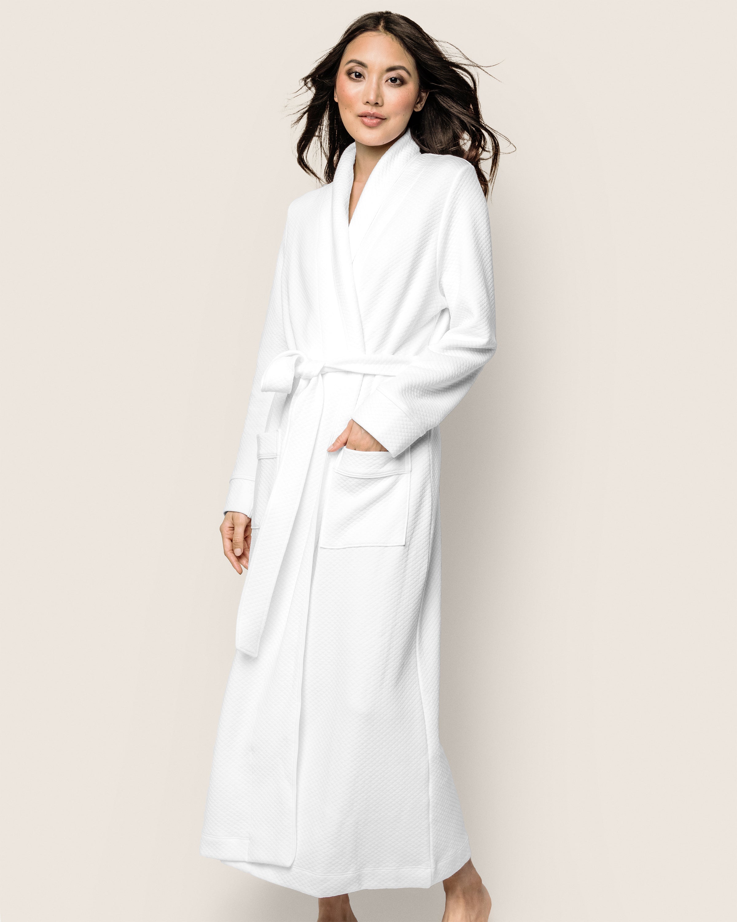 A person poses in a Petite Plume Womens Pima Ophelia Robe in White Jacquard, against a plain background. The robe boasts a luxury quilted effect and is made of Peruvian Pima cotton, featuring two pockets. Their hair flows gently as they smile slightly.