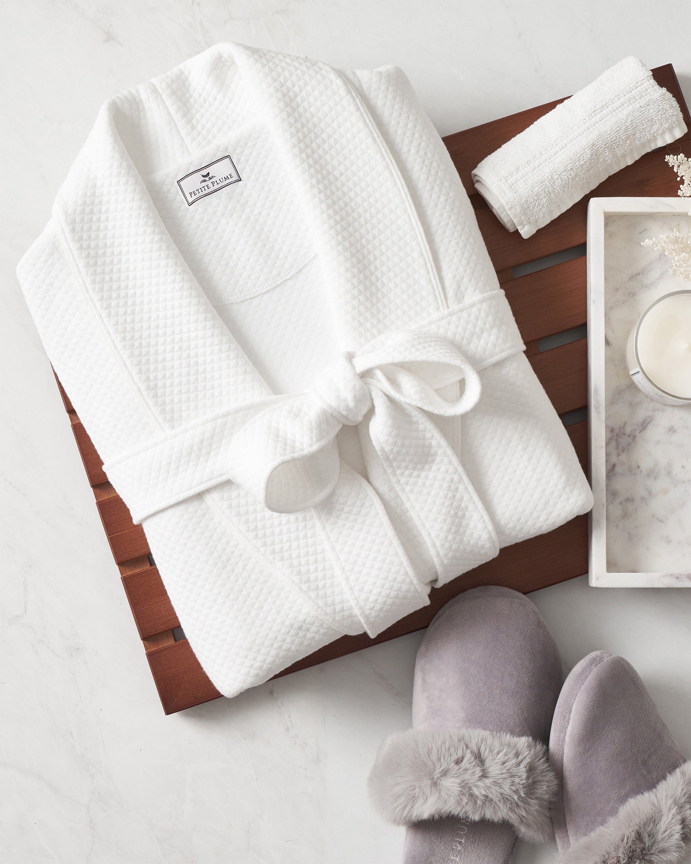 A folded Womens Pima Ophelia Robe in White Jacquard by Petite Plume sits on a wooden tray alongside gray fuzzy slippers, a rolled towel, a lit candle, and white flowers on pristine white marble.