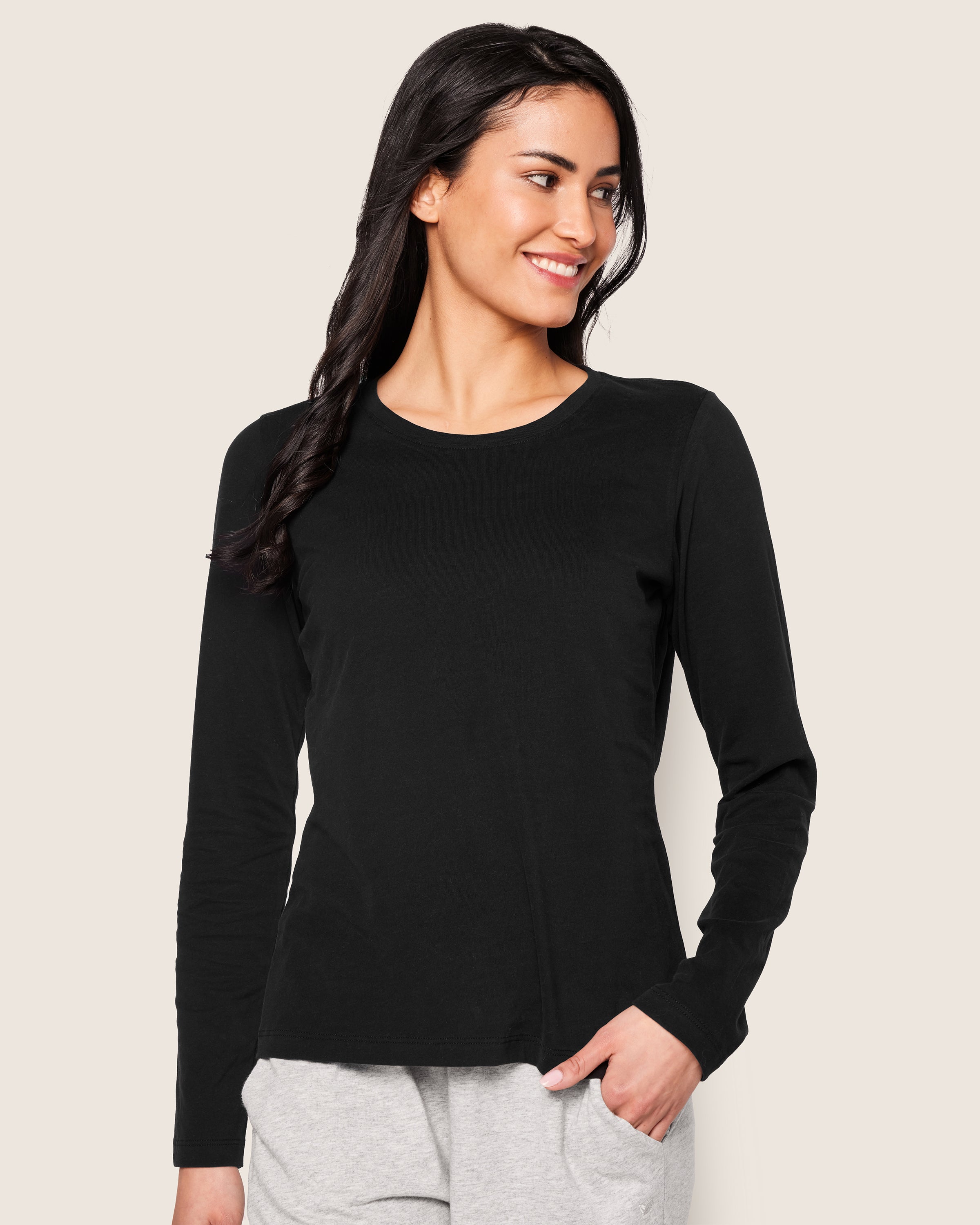A smiling woman with long dark hair wears a Petite Plume Womens Pima Long Sleeve Top in Black and light gray jersey knit pants, standing against a plain light background with one hand in her pants pocket.
