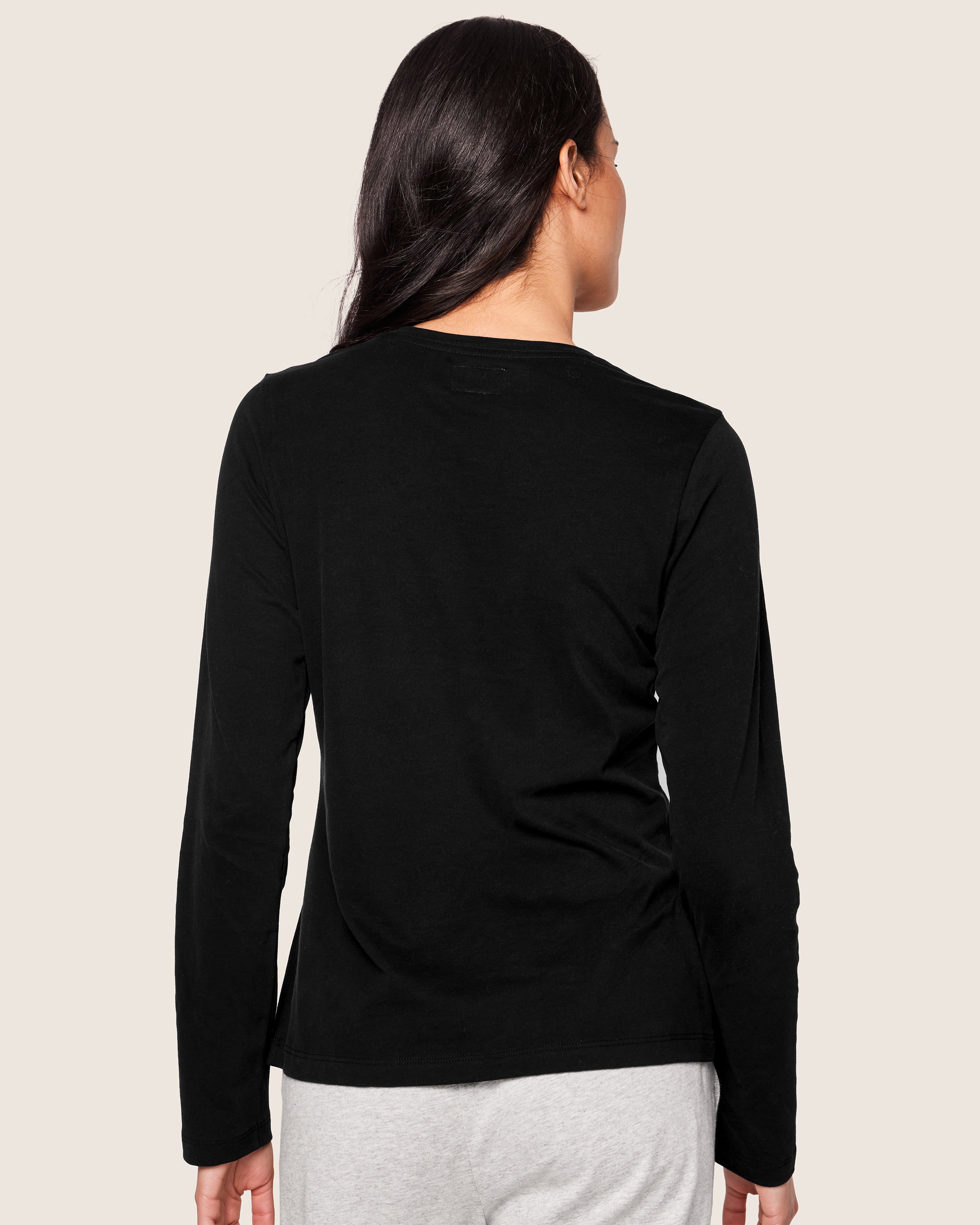 Seen from behind, a person with long black hair wears luxury attire: a Womens Pima Long Sleeve Top in Black by Petite Plume, paired with gray pants. The background is plain off-white.