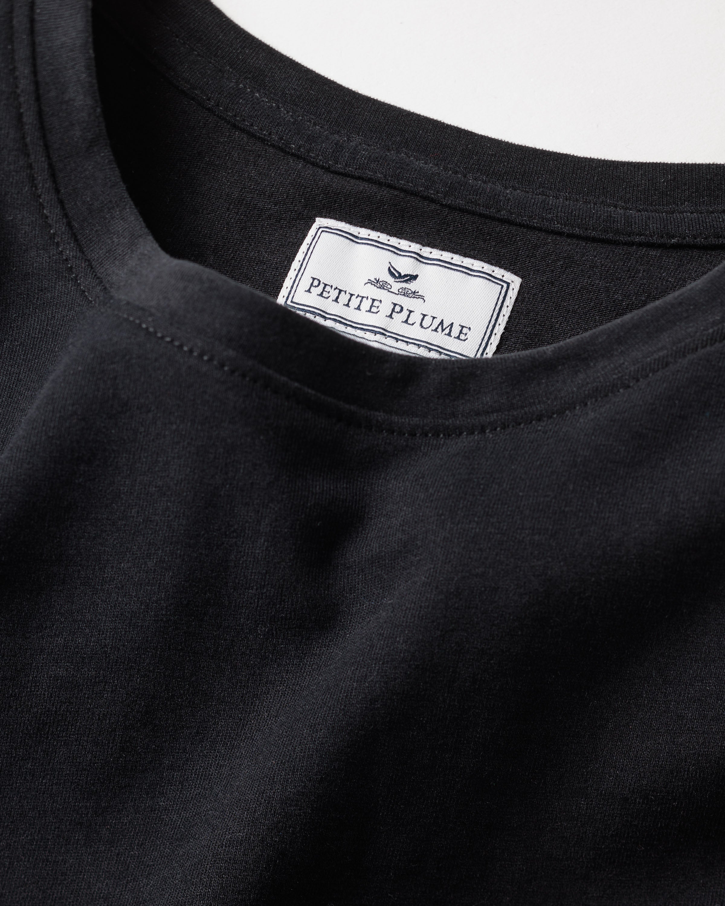 Close-up of Petite Plumes Womens Pima Long Sleeve Top in Black. The label inside, with a feather logo, reveals the brand. Made from soft jersey knit with visible Pima Cotton stitching along the neckline, it hints at luxury pajama quality.