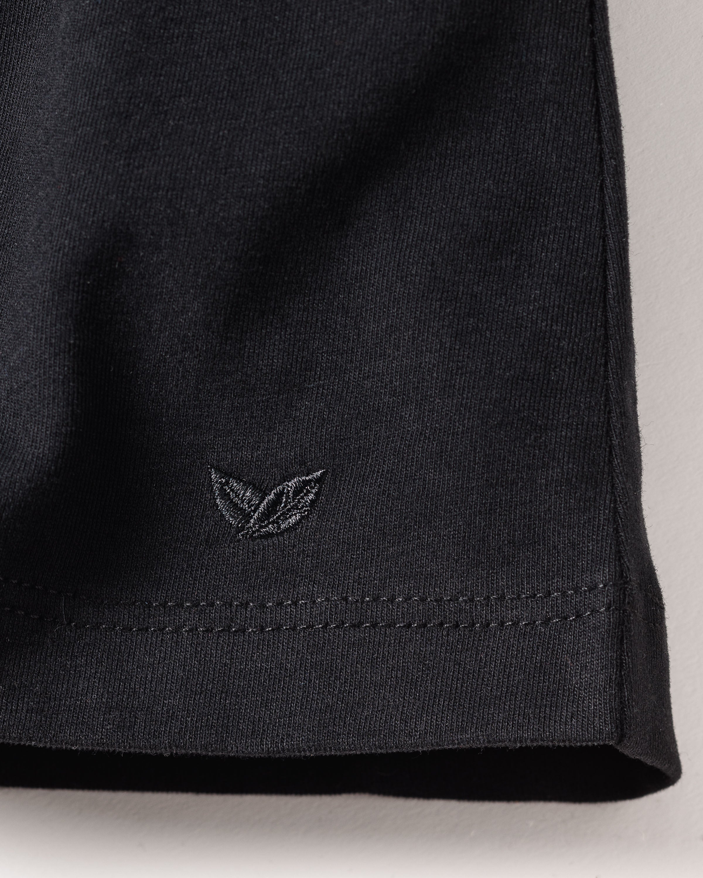 Close-up of the hem of a Womens Pima Long Sleeve Top in Black from Petite Plume, made from smooth jersey knit with a small black embroidered logo of overlapping leaves near the bottom. The fabric is as soft and neatly stitched as luxury pajamas.
