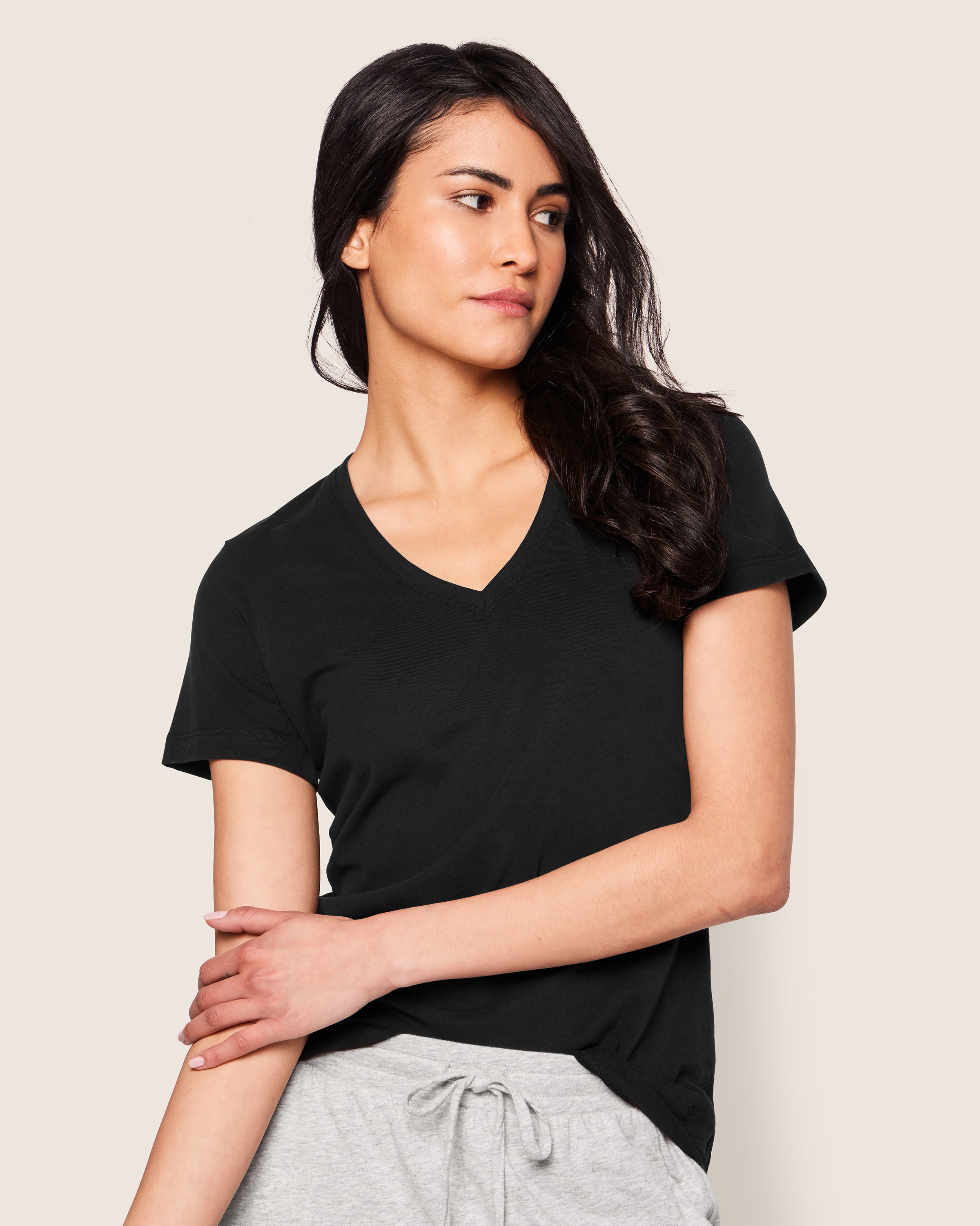 A woman in a Petite Plume Womens Pima Short Sleeve Top in black stands against a plain background, wearing gray drawstring pants. With long dark hair, she looks to the side with one arm crossed and the other resting on her forearm.