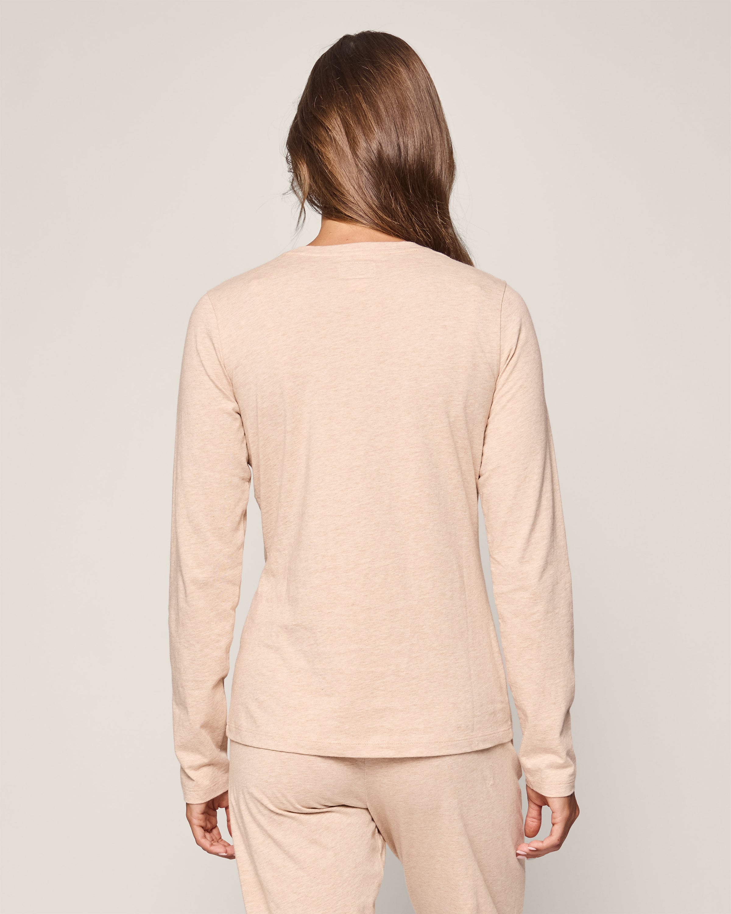 A person with long brown hair stands facing away in luxurious Petite Plume pajamas made from Peruvian Pima cotton, wearing the Womens Pima Long Sleeve Top in Camel and matching pants. The plain, light-colored background accentuates the fabrics elegance.