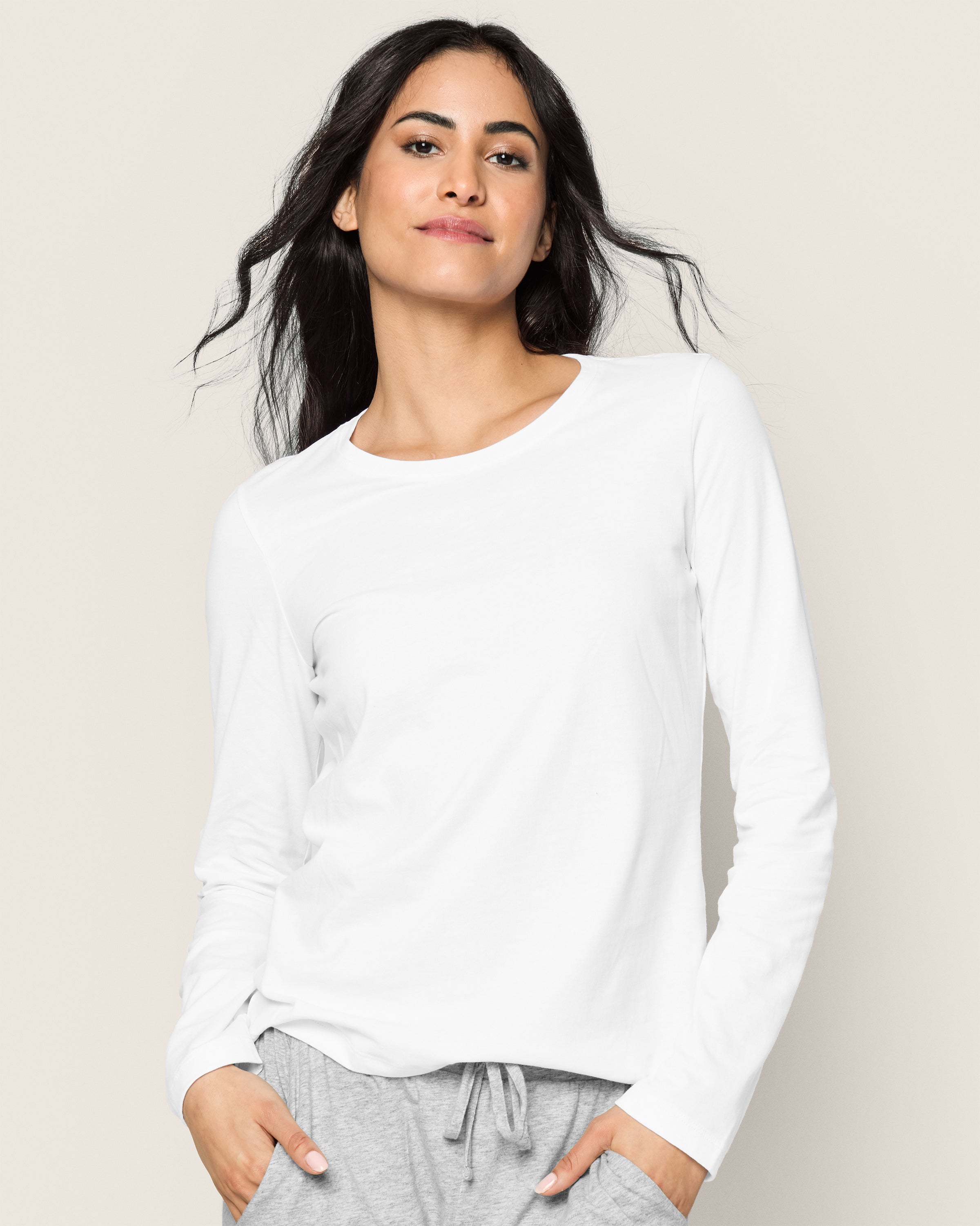 A person with long, dark hair wearing the Womens Pima Long Sleeve Top in White by Petite Plume and gray pants smiles slightly at the camera. The plain, light background enhances the scenes elegance.