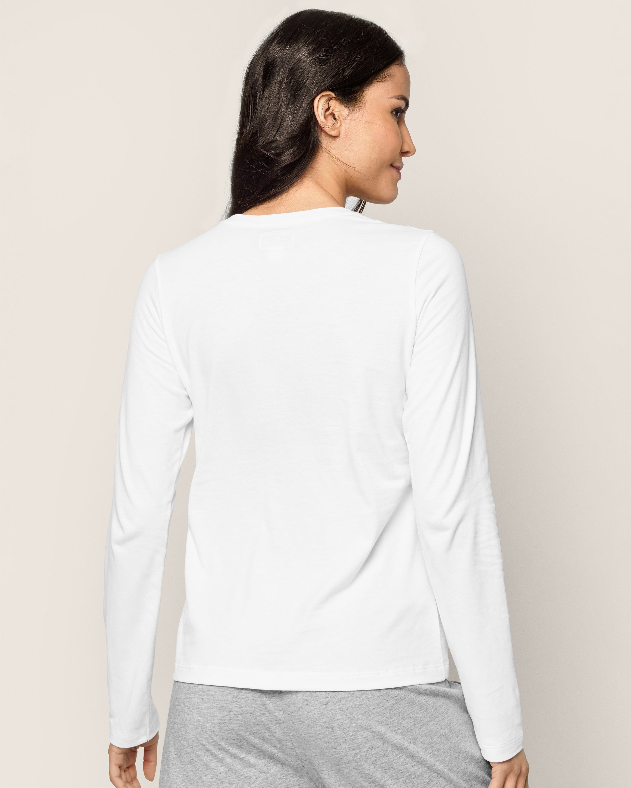A person with long dark hair stands against a beige background, seen from the back wearing Petite Plumes Womens Pima Long Sleeve Top in White, paired with gray pants made from luxurious Peruvian Pima cotton.