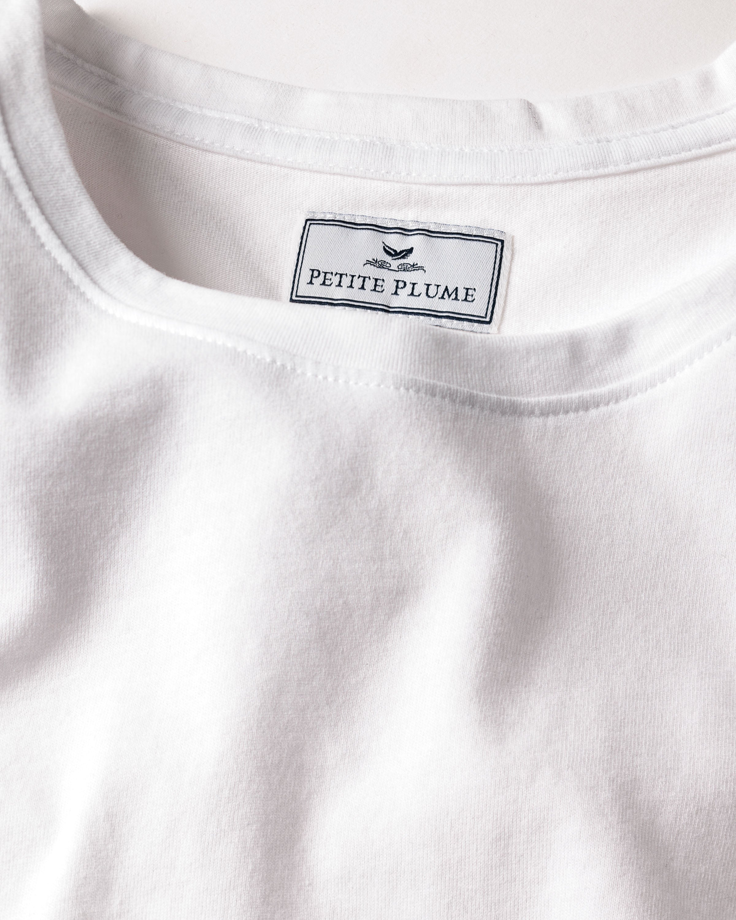 Close-up of a Petite Plume label with a bird logo sewn inside the neckline of a Womens Pima Long Sleeve Top in White, conveying the soft, smooth allure of luxury pajamas for serene nights.