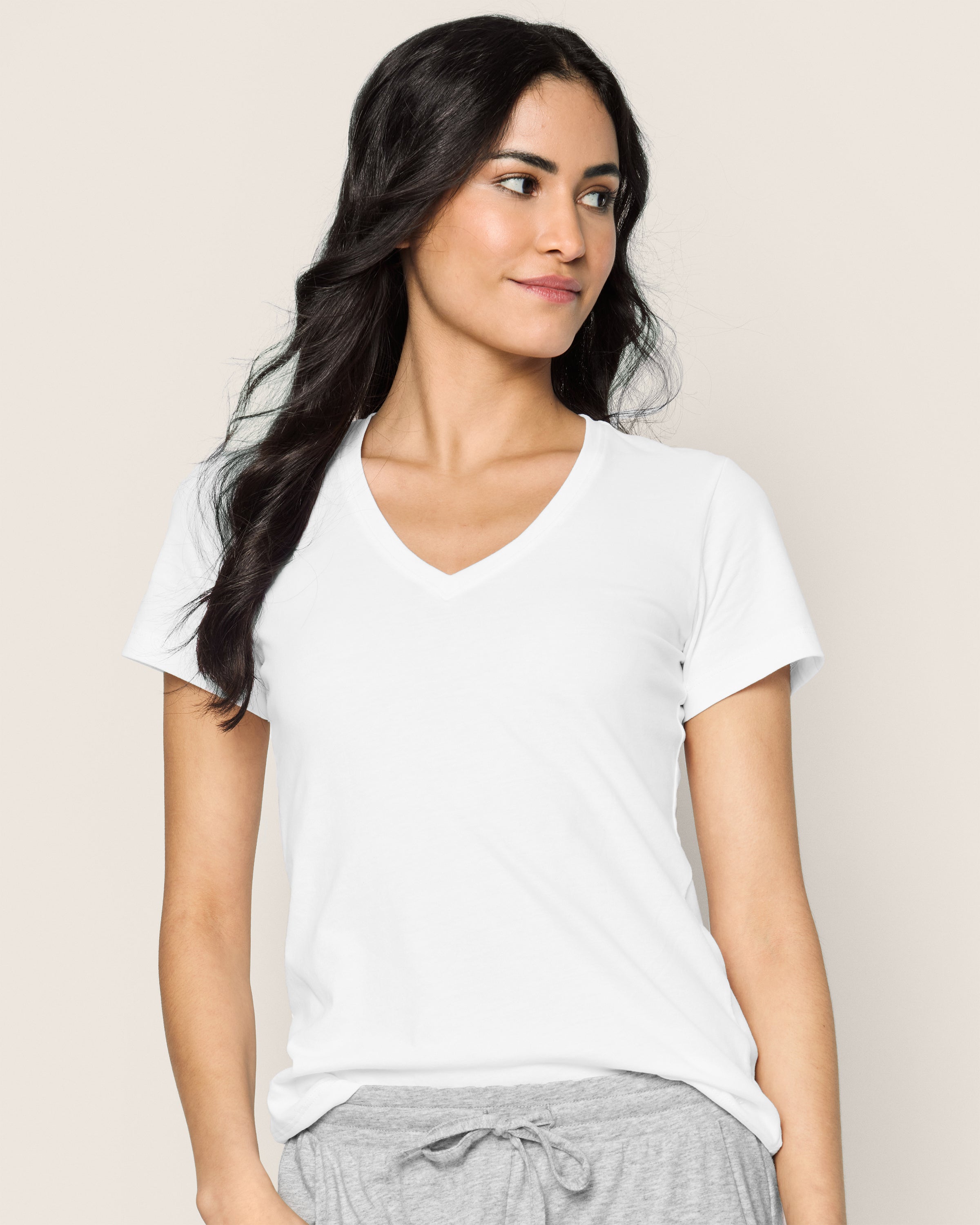 A woman with long dark hair is wearing a white Womens Pima Short Sleeve Top by Petite Plume and gray drawstring pants. She stands against a plain light-colored background, looking to her left with a slight smile.