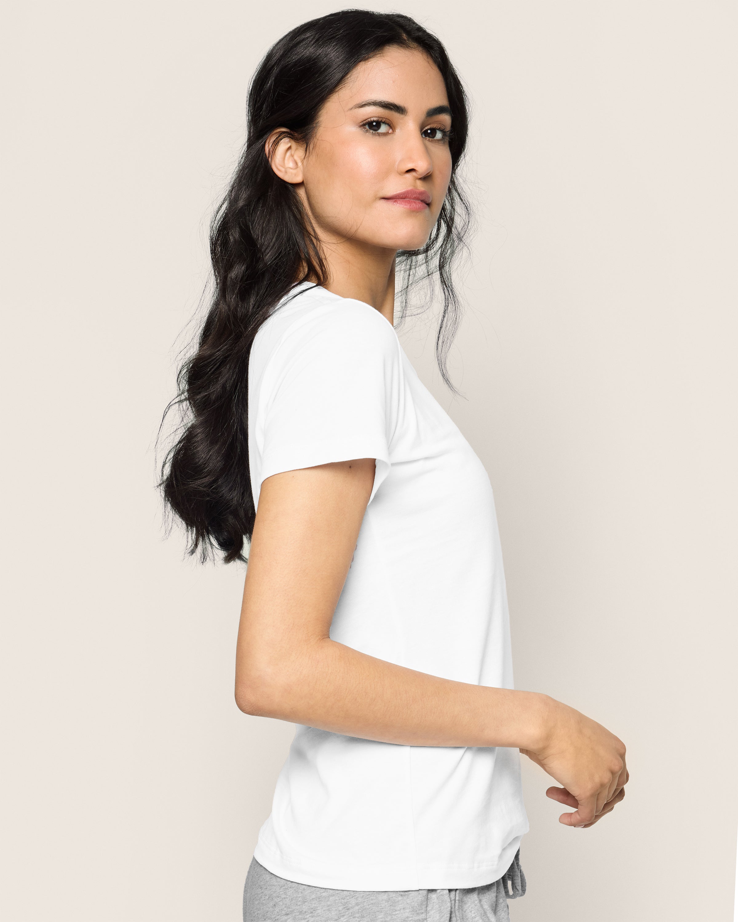 A person with long dark hair, dressed in a Womens Pima Short Sleeve Top in White by Petite Plume and gray soft jersey knit pants, stands against a plain light background, looking over their shoulder.