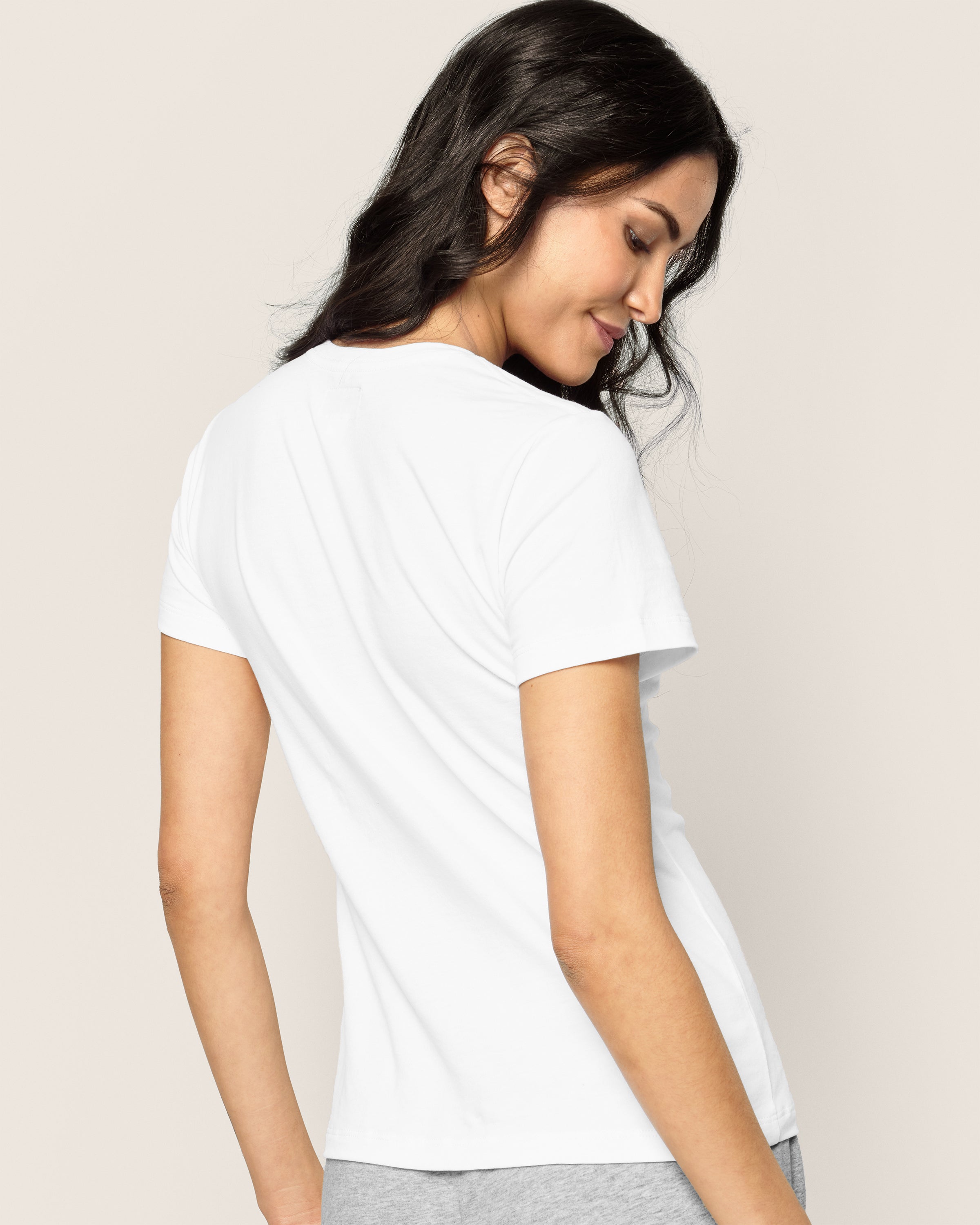 A person with long dark hair is smiling and looking over their shoulder, wearing Petite Plumes Womens Pima Short Sleeve Top in White and gray jersey knit pants against a plain light background.