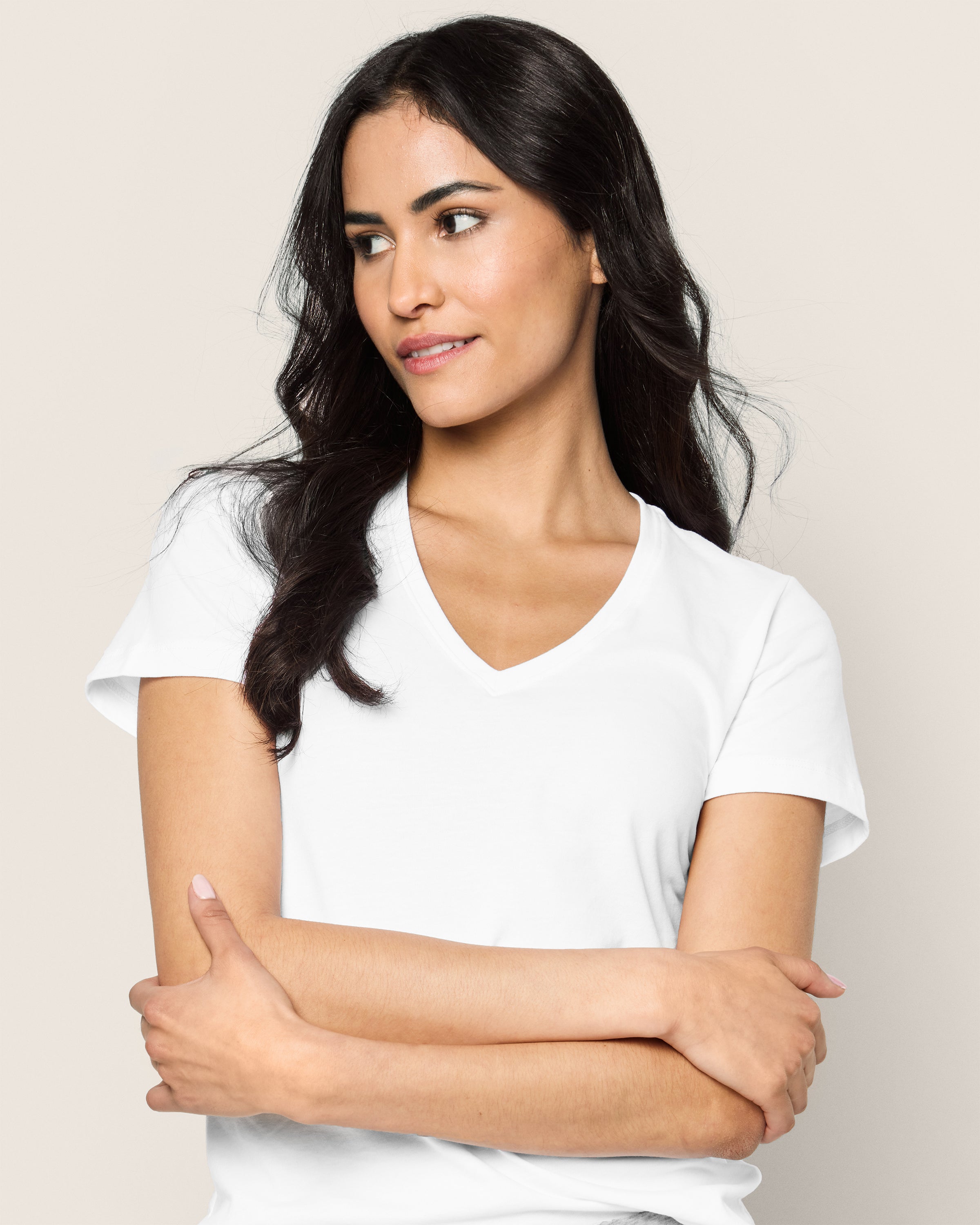 A person with long dark hair wears a Womens Pima Short Sleeve Top in White by Petite Plume. They cross their arms and look to the side with a soft smile against a light neutral background, embodying the comfort and elegance akin to luxury pajamas.