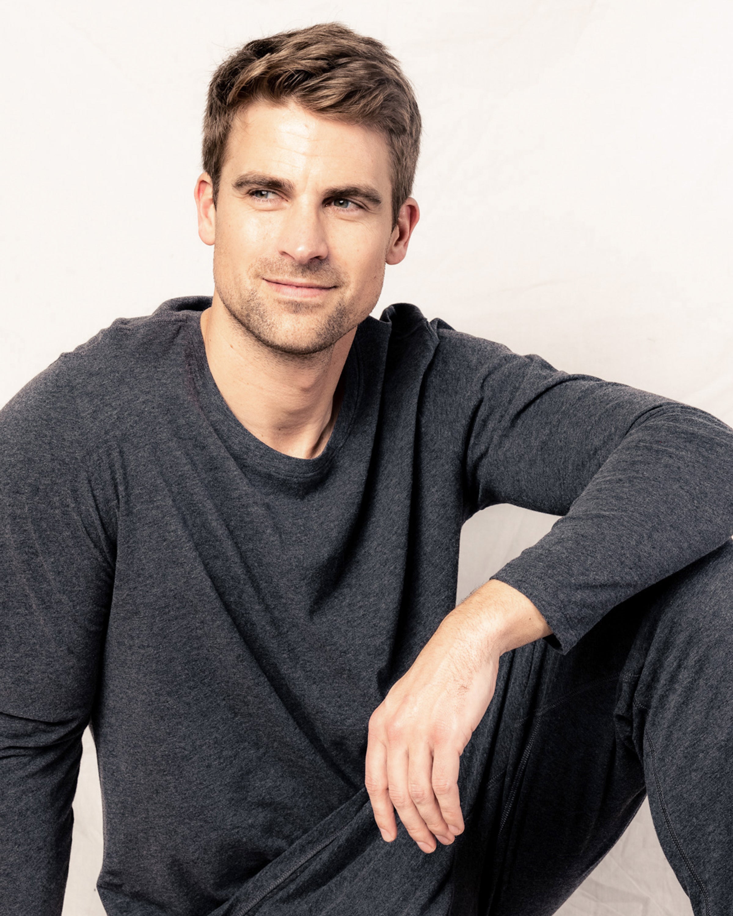 A man with short brown hair sits casually against a light background, dressed in a Petite Plume Mens Pima Crew Neck Long Sleeve Top in Dark Heather Grey. Made from soft Peruvian Pima cotton, it hints at the comfort of jersey knit pajamas. His right arm rests on his knee as he gazes thoughtfully to the side.