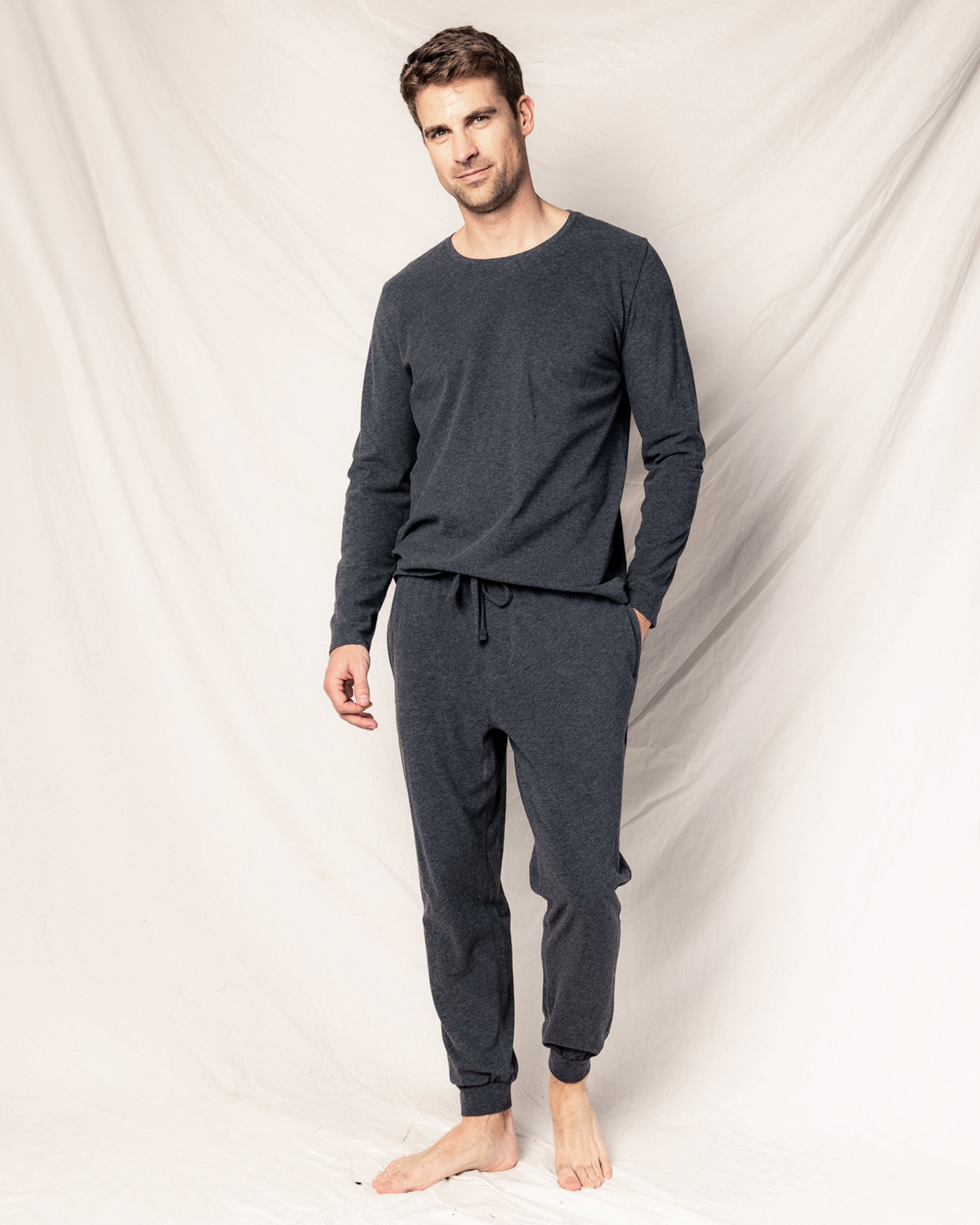 A person stands barefoot against a light beige backdrop, wearing the Mens Pima Crew Neck Long Sleeve Top in Dark Heather Grey by Petite Plume. Crafted from soft Peruvian Pima cotton, the breathable jersey knit pajamas appear comfortable and relaxed, with hands casually placed in pockets.