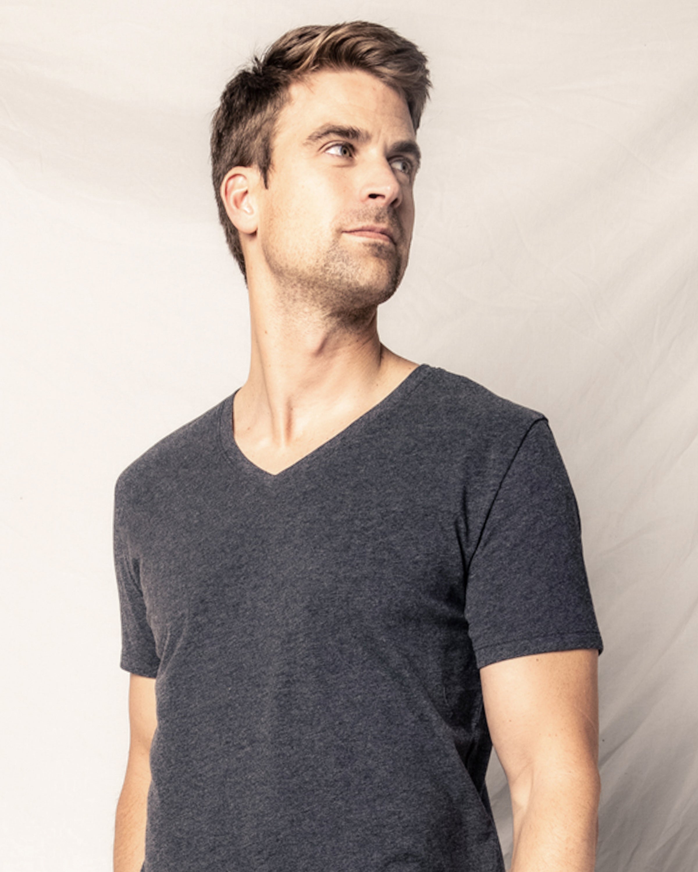 A man in a Mens Pima V-Neck Top in Dark Heather Grey by Petite Plume stands against a light beige backdrop, looking to the side.