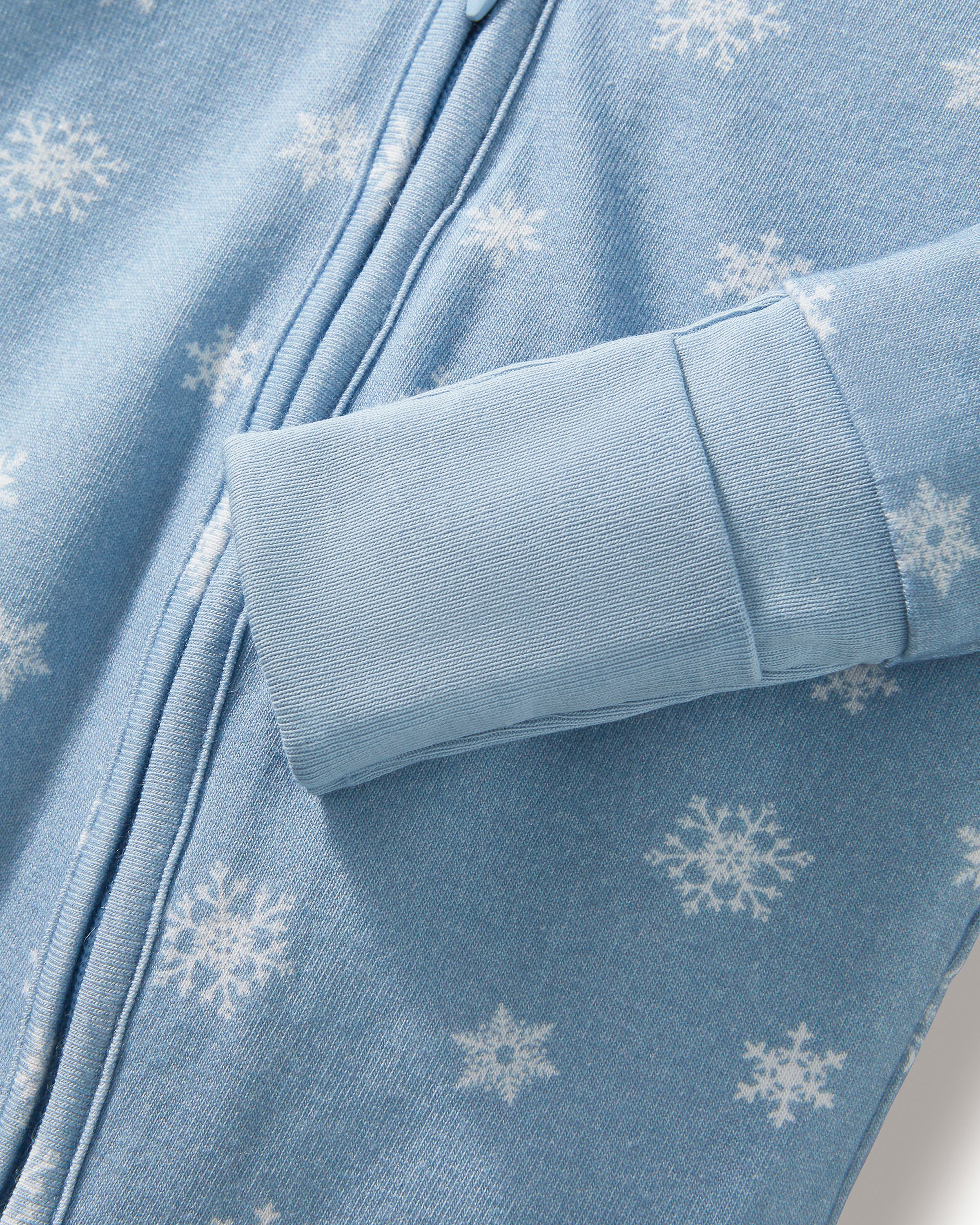 The Baby's Pima Peekaboo Romper in Periwinkle Wonderland features a light blue snowflake pattern, long sleeves with fold-over cuffs, and a diagonal zipper from neckline to left ankle; one sleeve and one leg are folded.