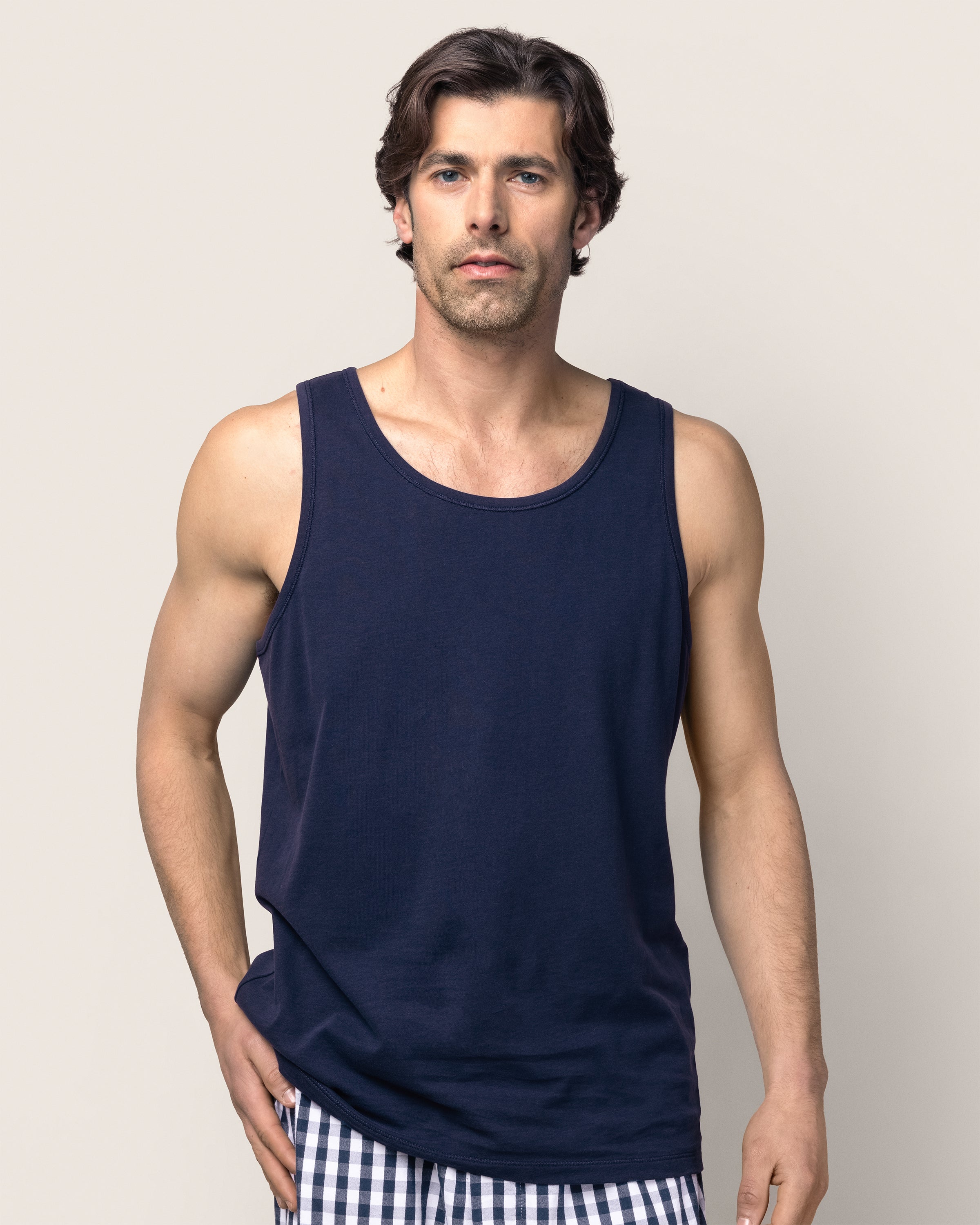 A man with medium-length wavy hair wears a Petite Plume Mens Pima Tank Top in Navy and Peruvian Pima cotton checkered shorts. Against a light gray background, he looks at the camera with a neutral expression, right hand on his hip, evoking a relaxed bedtime ritual vibe.