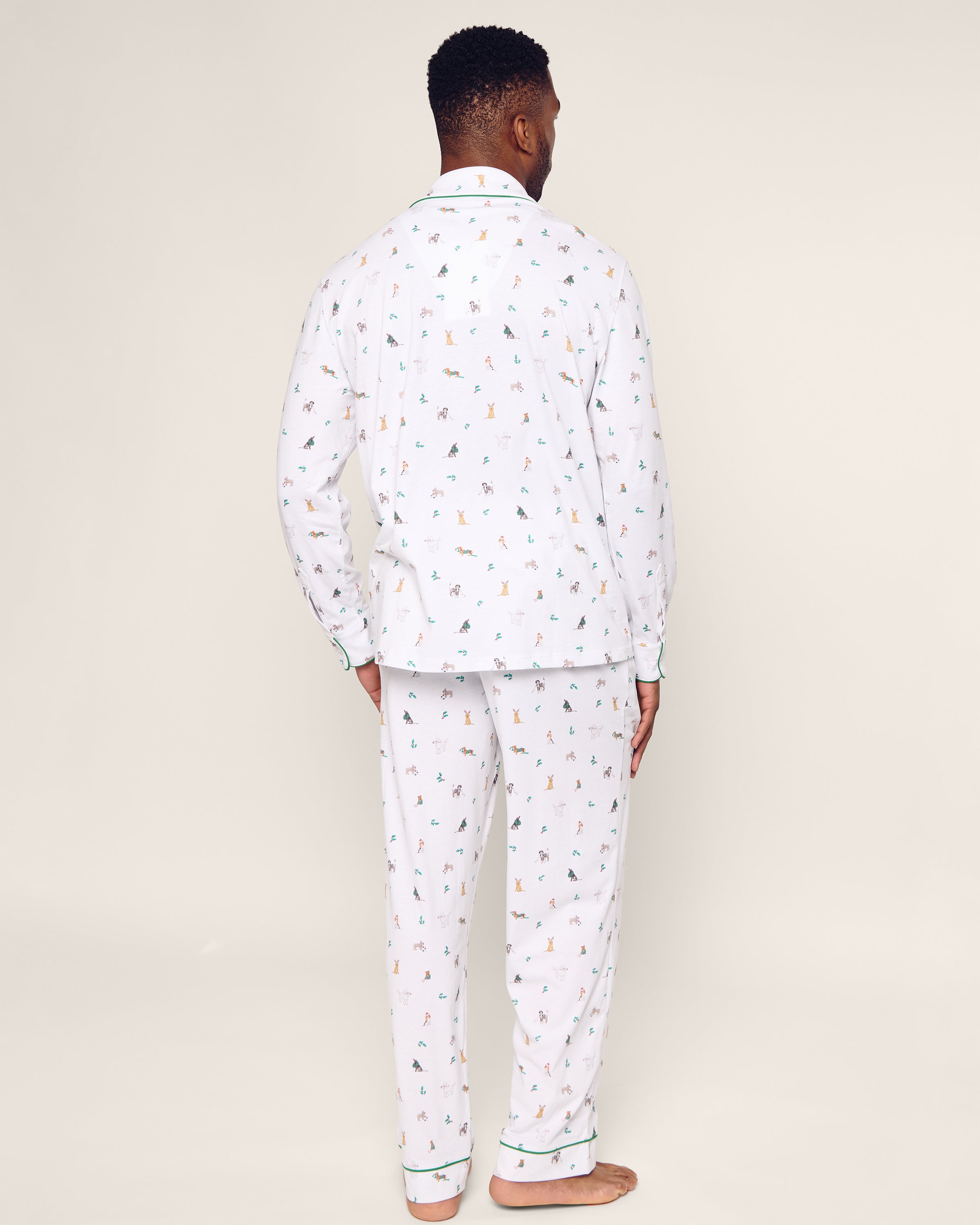 Rear view of a person in the Petite Plume Mens Pima Pajama Set in Jingle Paws, standing on a beige floor. Featuring long sleeves and full-length pants, its crafted from soft Peruvian Pima cotton with small colorful prints. Ideal holiday sleepwear, their hands rest by their sides.