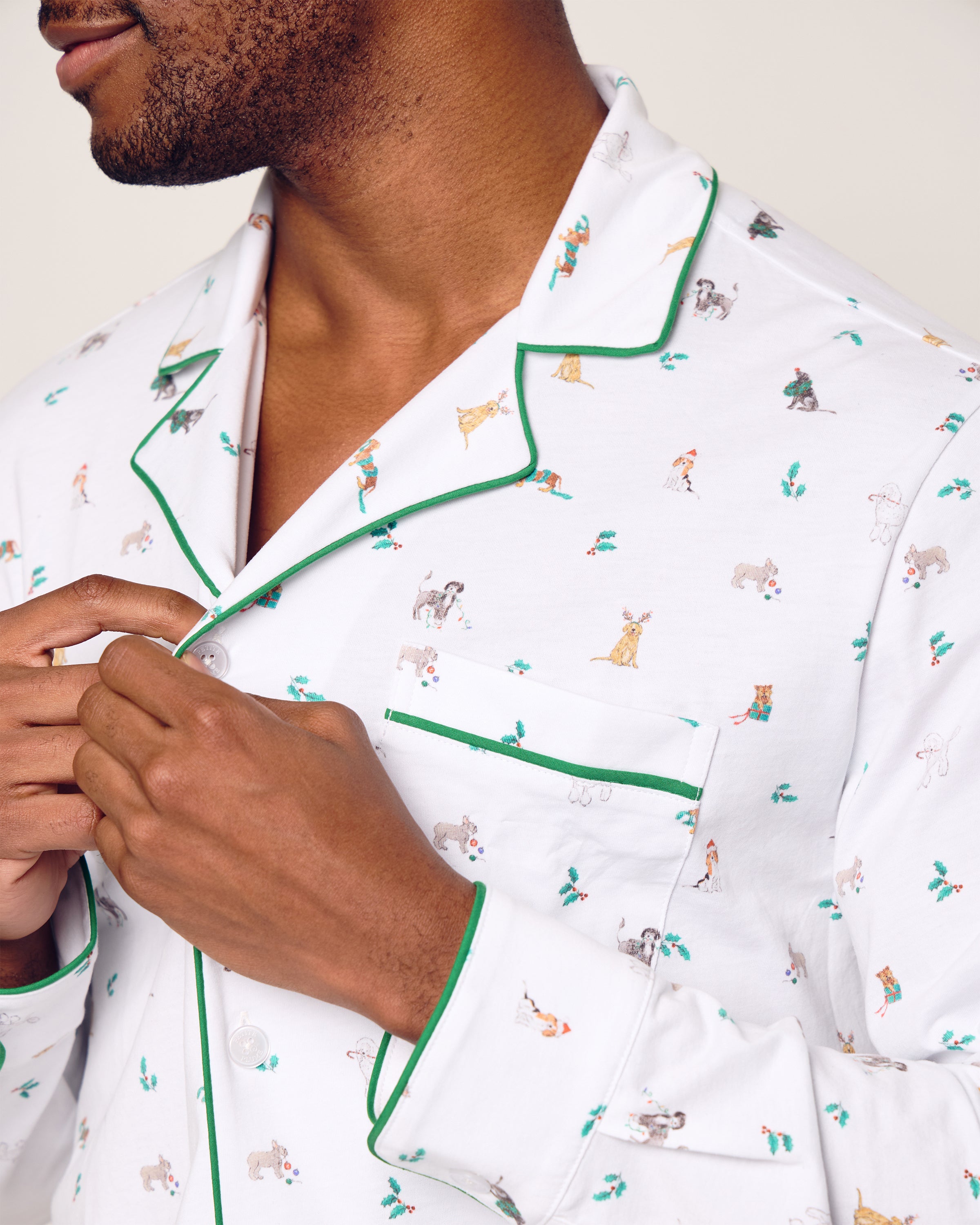 A man buttons the top of his Petite Plume Mens Pima Pajama Set in Jingle Paws, made from soft Peruvian Pima cotton. The white pajamas with green piping feature playful animal patterns, adding a whimsical touch against a plain, neutral background.
