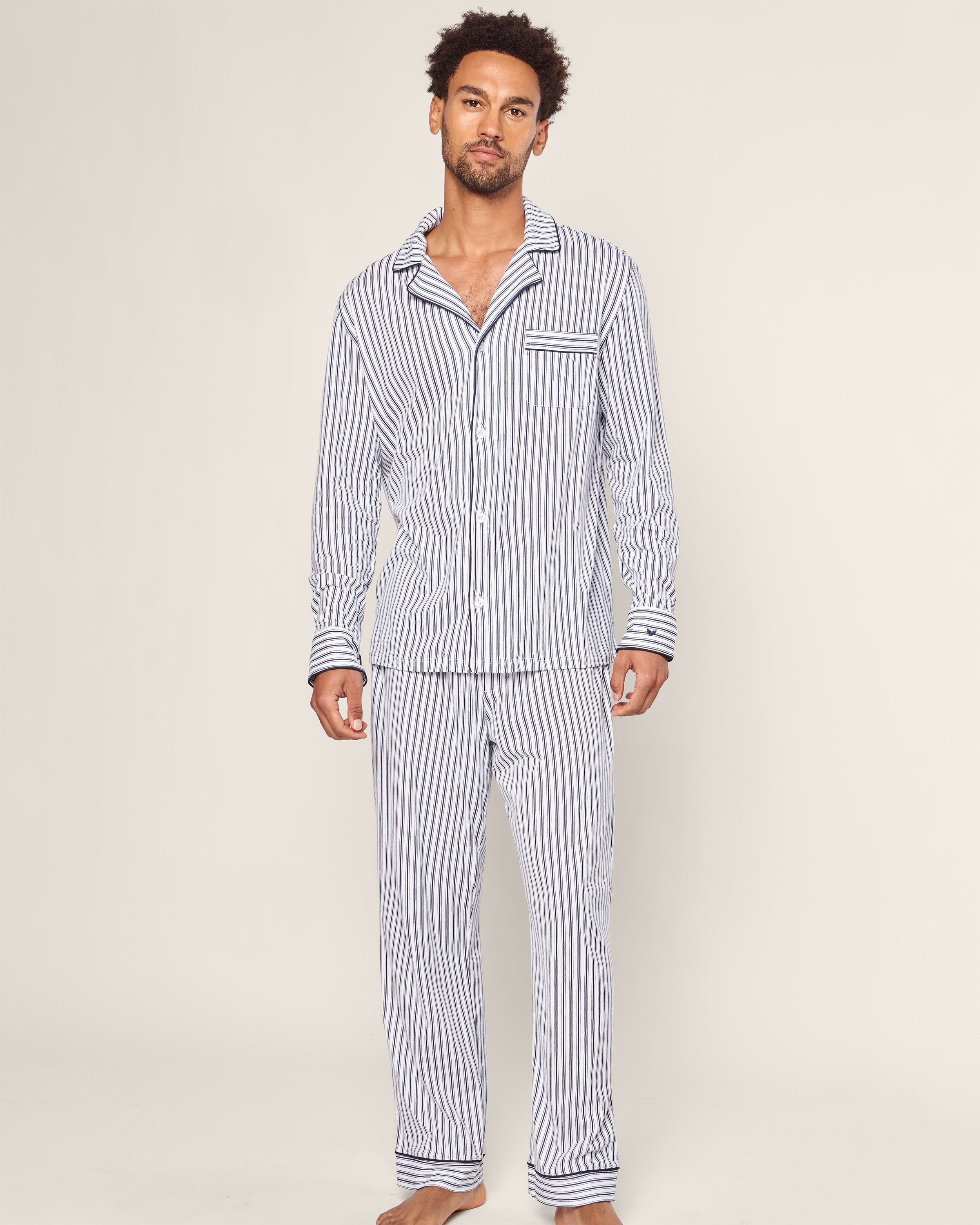 A man dons Petite Plumes Mens Pima Pajama Set in Navy French Ticking. This classic, long-sleeved button-up shirt and matching pants crafted from soft Pima cotton exude luxury and style as he stands barefoot against a plain light background.
