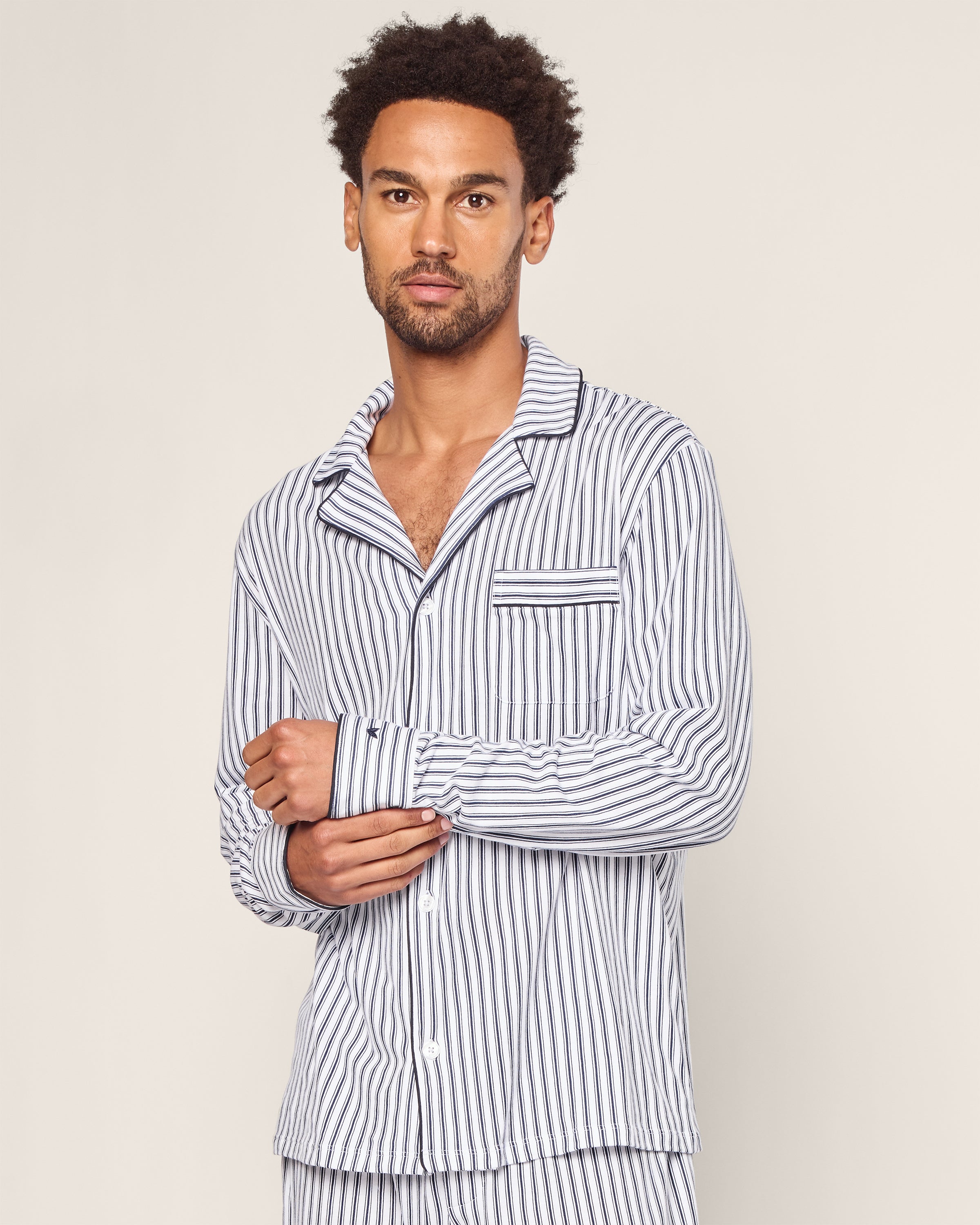 A person with curly hair wears Petite Plumes Mens Pima Pajama Set in Navy French Ticking, showcasing a luxurious blue-and-white striped design. The classic top has a collar and chest pocket, and the plain, light background accentuates the elegance of this premium sleepwear.