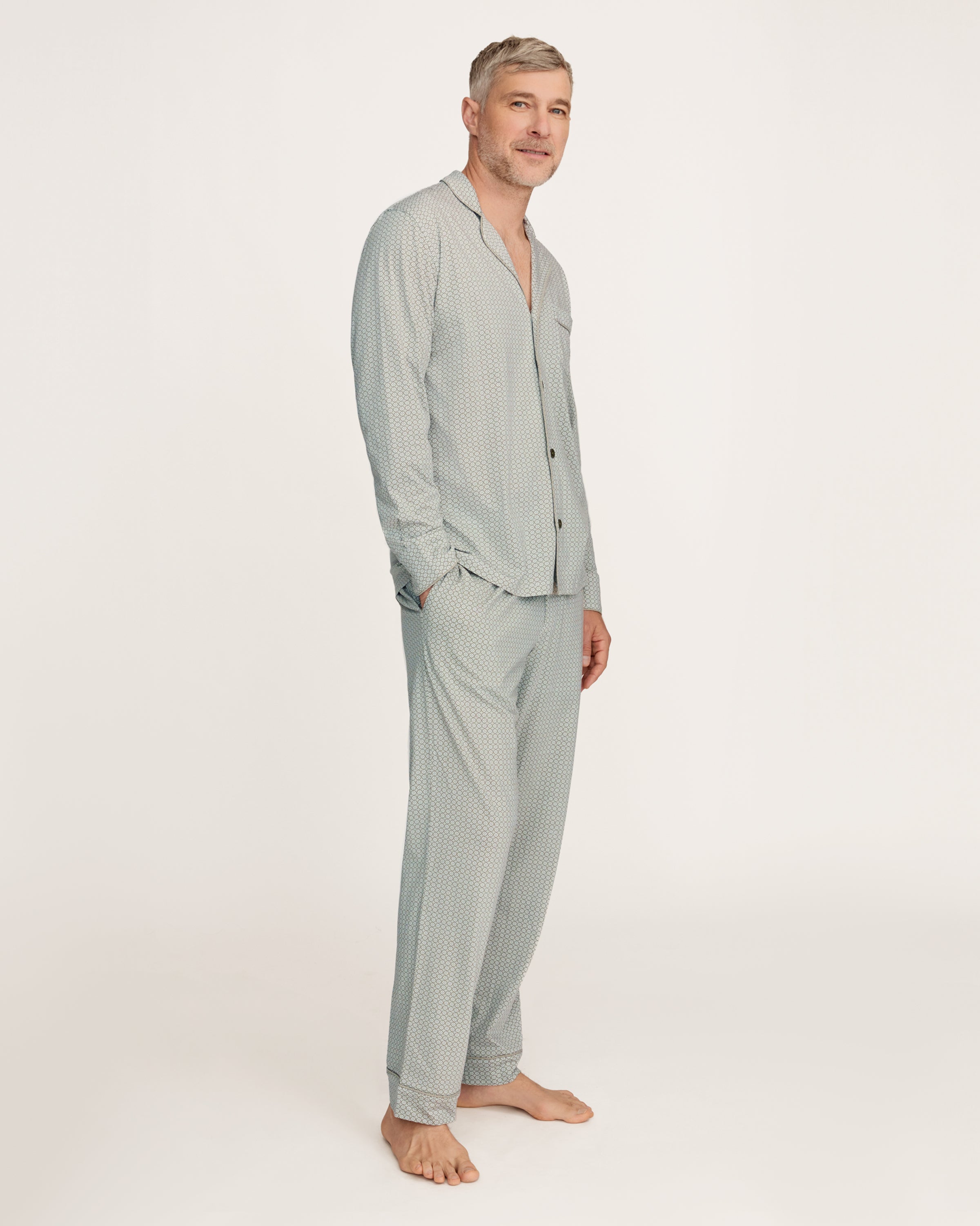 A man with short gray hair and light stubble stands barefoot in the Men's Lightweight Pima Pajama Set in Sage Geo against a white background, gazing to the side with a relaxed expression.