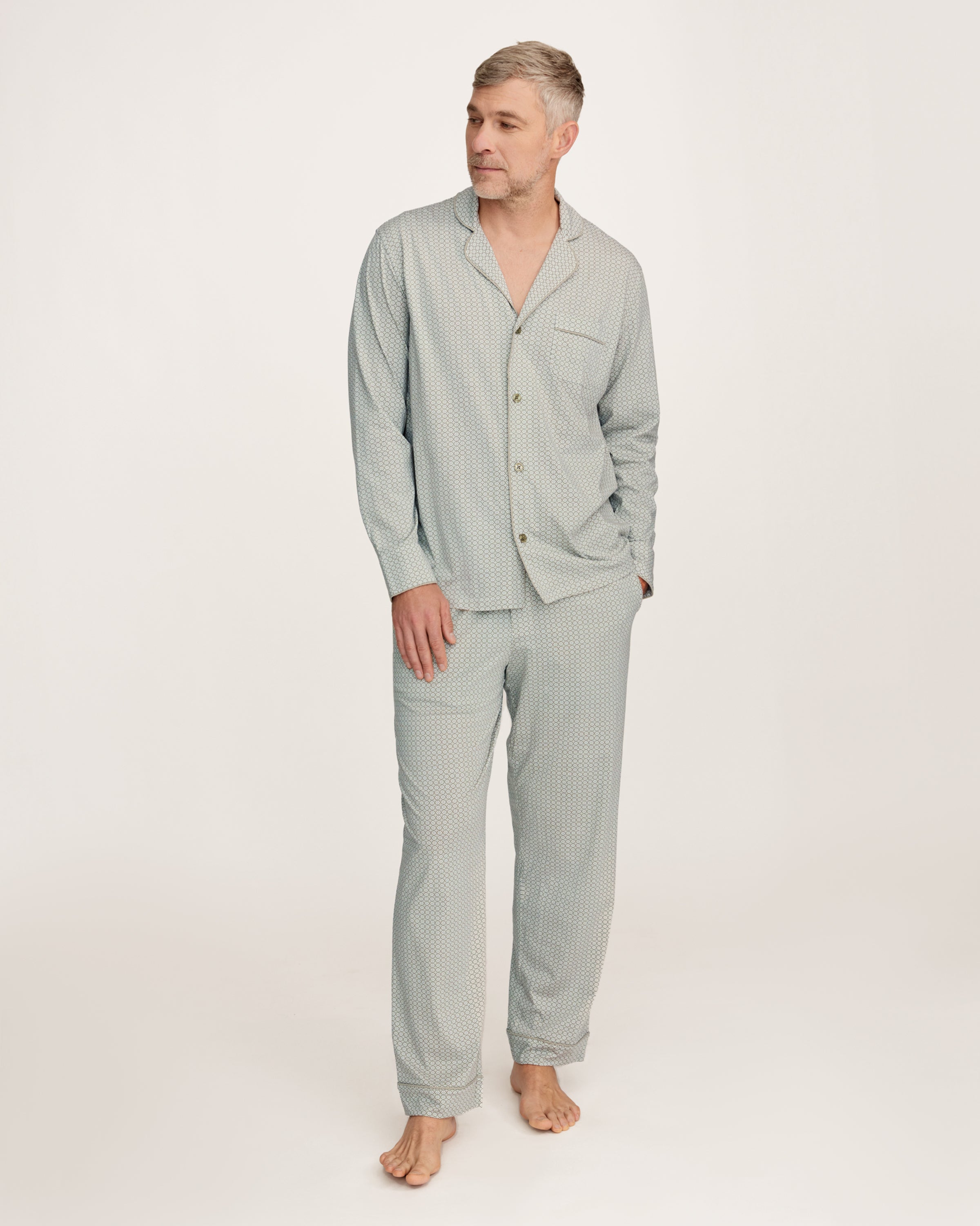 A man with short gray hair and light stubble stands barefoot in the Men's Lightweight Pima Pajama Set in Sage Geo against a white background, gazing to the side with a relaxed expression.
