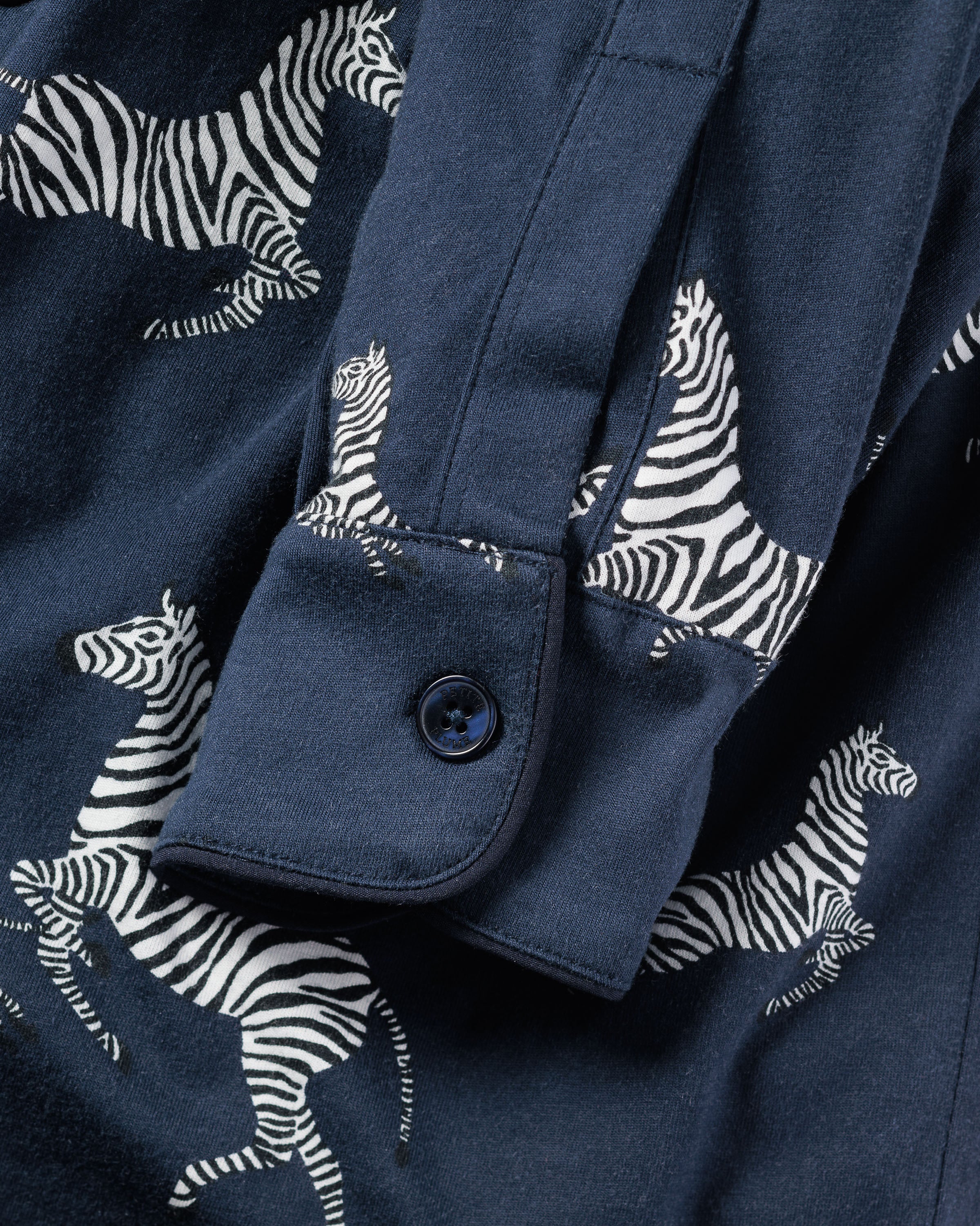 A close-up of the Mens Pima Pajama Set in Zèbre de Nuit by Petite Plume showcases their luxury sleepwear with an intricate white zebra pattern on navy blue fabric, highlighting a buttoned cuff on the sleeve and capturing the elegance and detail of this Pima cotton piece.